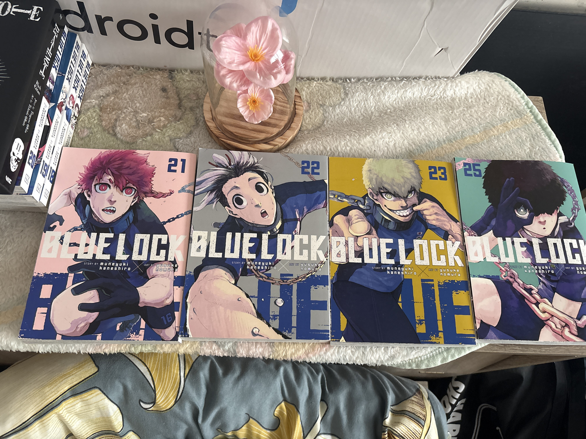 The image displays a stack of manga volumes, predominantly from the "Blue Lock" series, arranged vertically. The manga covers feature distinct character art and volume numbers. From top to bottom, the visible volumes are numbered 16, 21, 22, 23, and 25. Below the manga, a dark t-shirt with white text is partially visible. The scene is set on a light-colored, textured surface, possibly a rug or blanket, with a patterned fabric to the left. A wooden coaster holding pink artificial flowers is also present to the right of the manga stack. The overall lighting suggests an indoor environment. There are no people or animals depicted in the image. The location of Burgas, Bulgaria is not visible or inferable from the provided image.