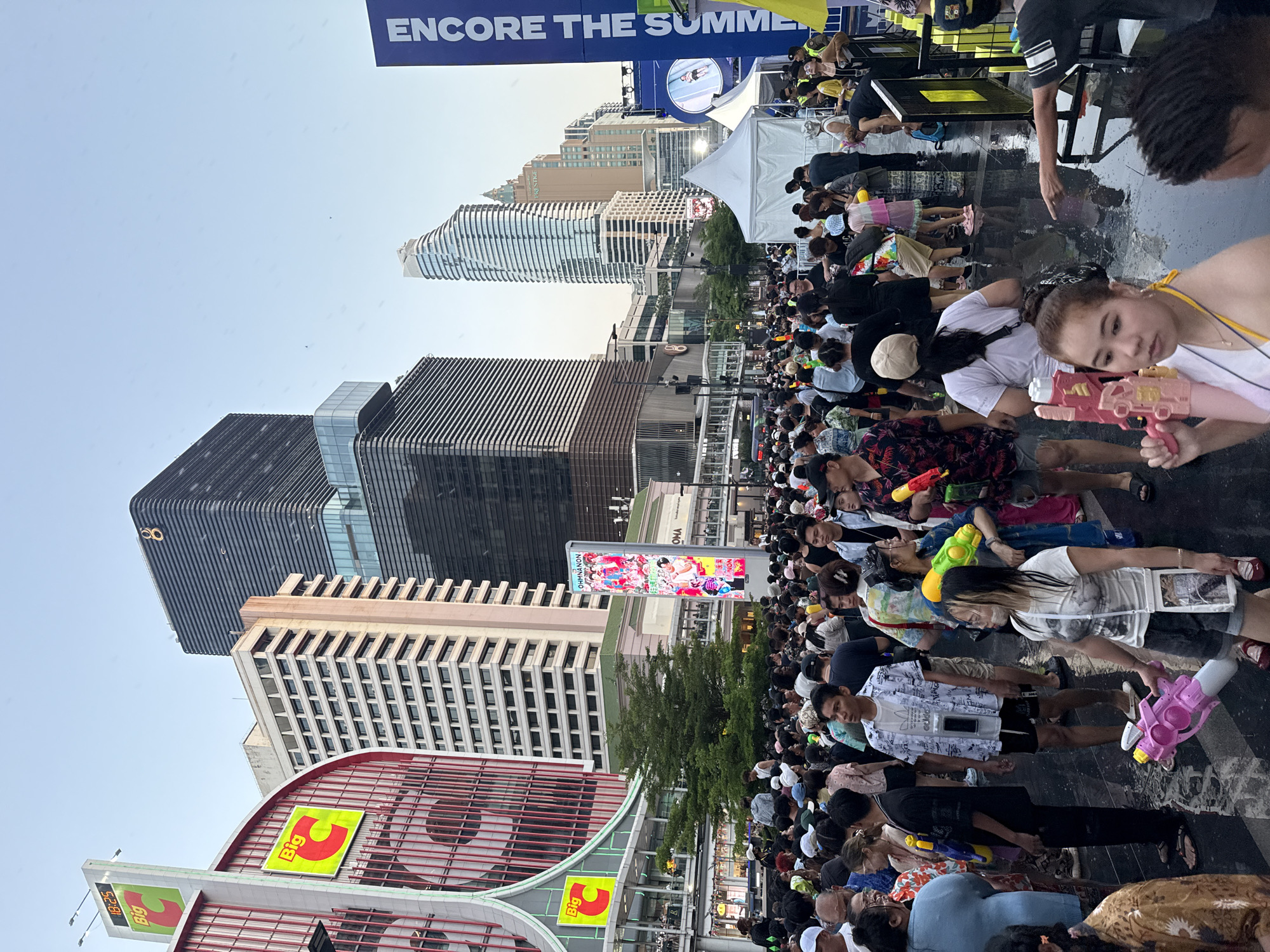 The image depicts a large crowd of people standing in a public space in Bangkok, Thailand, with modern buildings in the background. Many people are holding water guns. The ground appears wet. The signage for the Big C shopping center is visible, indicating the location. A banner advertising "ENCORE THE SUMMER" is also present.