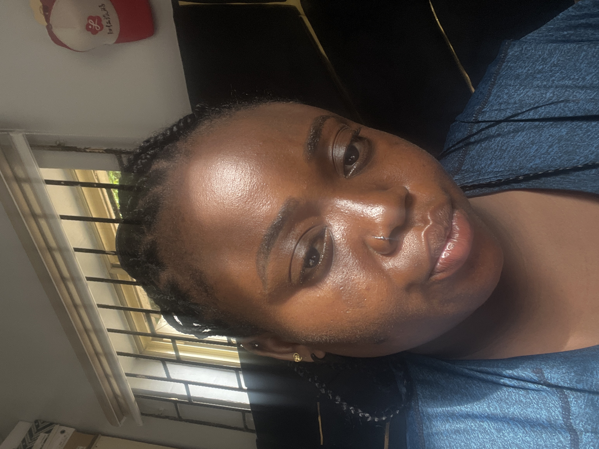 A portrait-style image, originally oriented horizontally but displayed vertically, captures a person with dark skin and braided hair, looking towards the camera. The subject's face is well-lit by natural light streaming in from a window to their left. Their skin appears dewy, reflecting the light, and they have a neutral expression with slightly parted, glossy lips. They are wearing a blue, textured top.
The setting is an indoor space, likely a room in Kubwa, Nigeria. A window with horizontal and vertical security bars is visible in the background, offering a glimpse of green foliage outside. Sunlight entering through this window illuminates parts of the subject's face and upper body. In the upper left corner of the original image, a small red and white decorative item, possibly a cap or ornament, is visible. This item prominently features the English word "Loves" followed by the Korean script "사랑해" (saranghae), which means "I love you." The remainder of the background is dark and out of focus. The scene suggests a casual, daytime portrait.