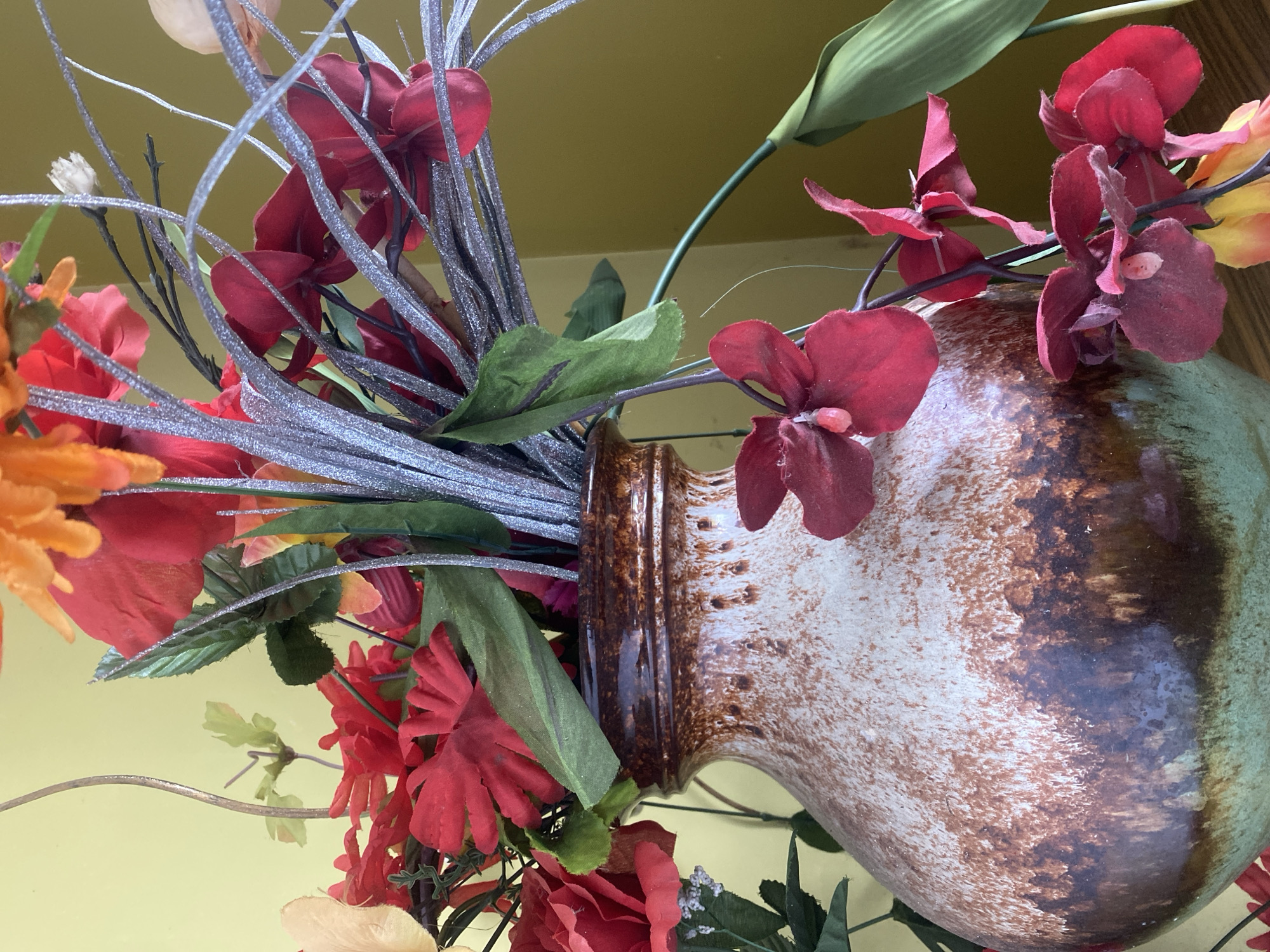 A close-up shot shows a vase filled with artificial flowers. The vase is speckled brown and green, with a bulbous shape and a narrow neck. The flowers are a mix of red, orange, and yellow, with some green foliage. The arrangement includes glittery silver twigs. The background is a plain yellow wall. The lighting is bright, suggesting an indoor setting, possibly daytime. There is no text visible in the image.