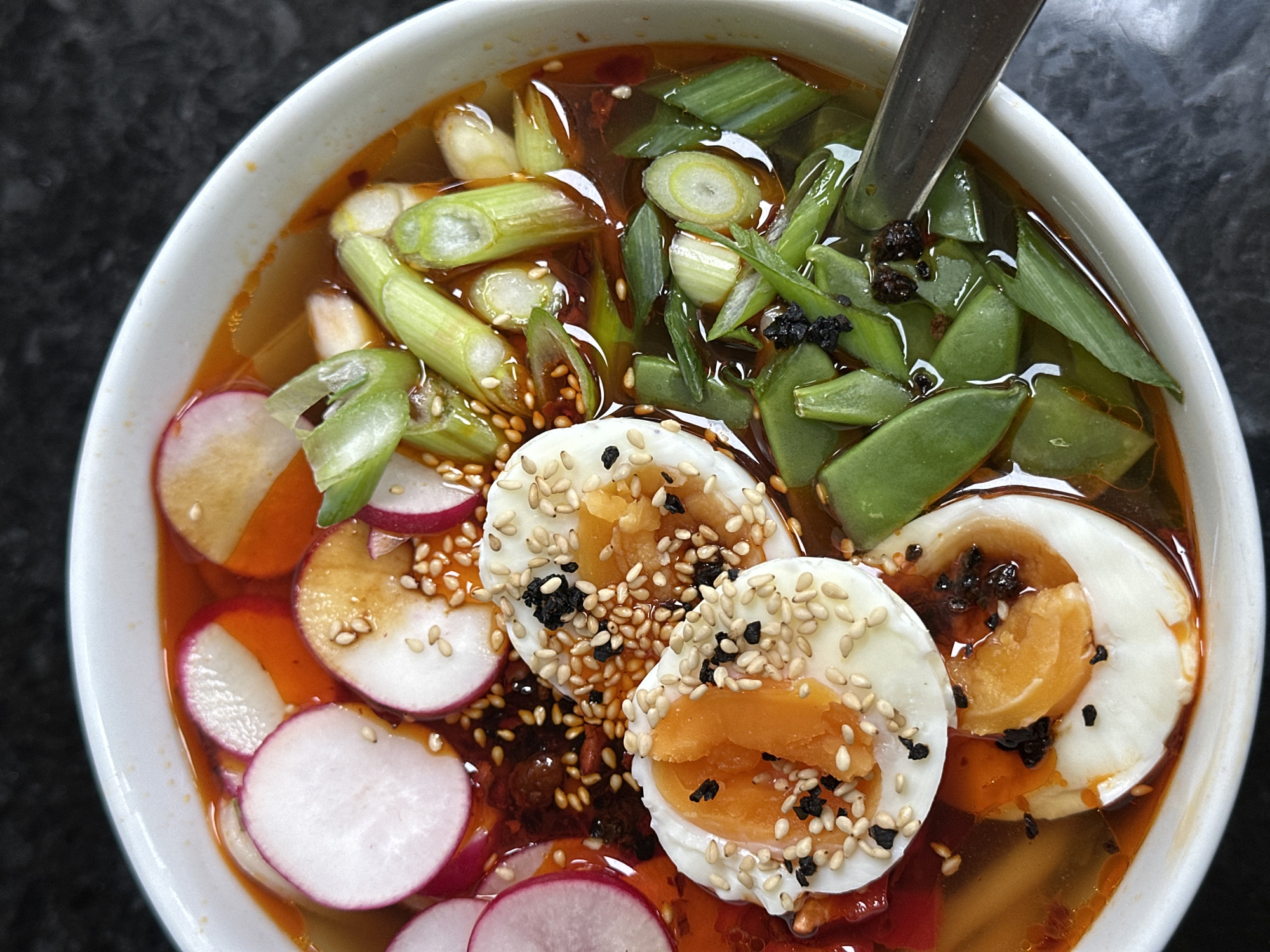 A white ceramic bowl is depicted from an overhead perspective, containing a soup or ramen dish. The soup features a reddish-orange broth with visible specks of chili oil. Ingredients include multiple circular slices of red and white radishes, abundant chopped green onions/scallions, and other green vegetable pieces, possibly snap peas. Centrally positioned are two halves of a soft-boiled egg, revealing firm white albumen and soft orange yolks, both generously topped with a mix of white and black sesame seeds. A silver-colored spoon is partially submerged in the broth in the lower right quadrant of the bowl. The background is a dark, out-of-focus surface. The image offers no visual information to confirm a location within the city of Wealden, United Kingdom.