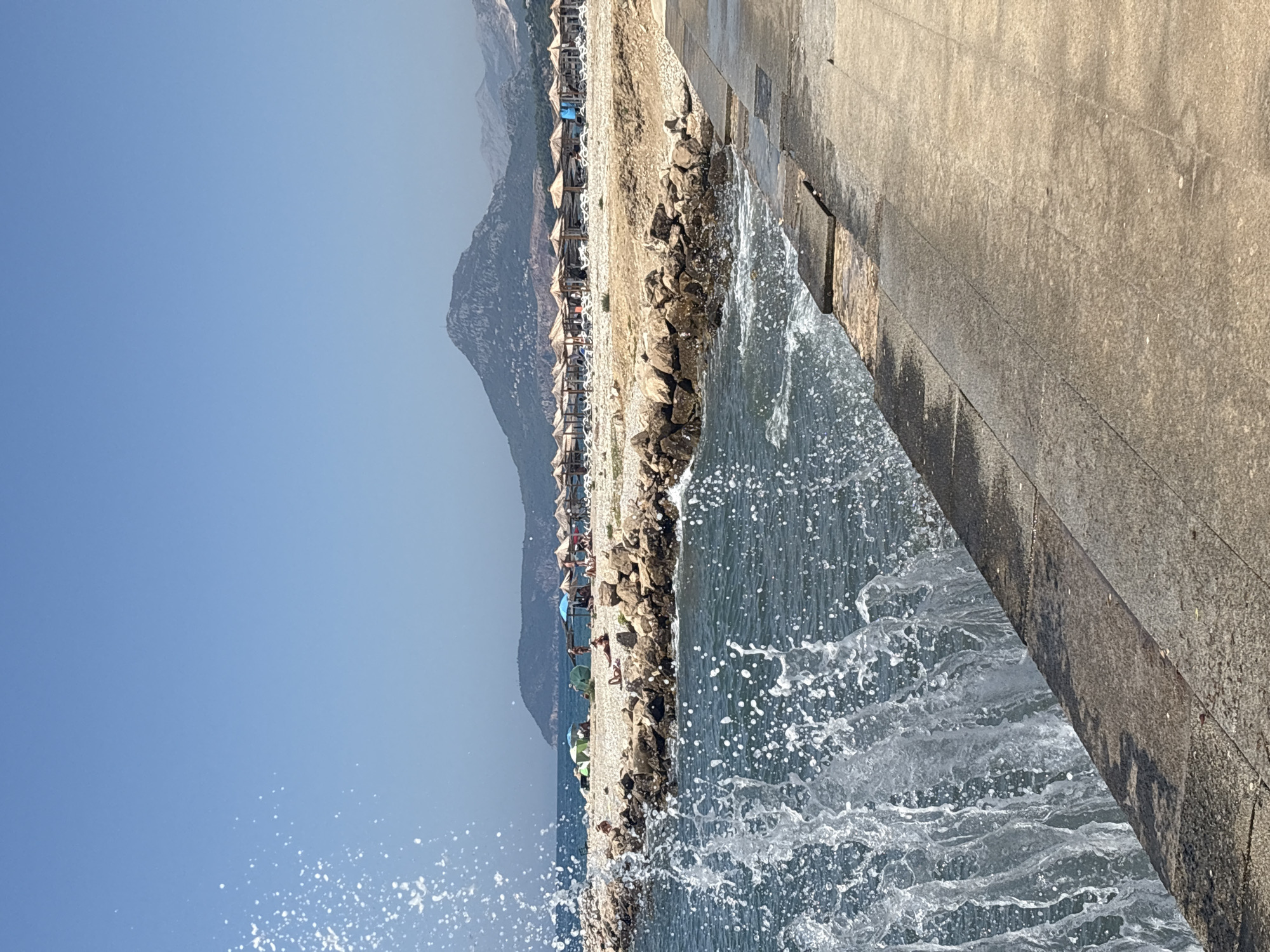 This is a photograph taken at a beach in Bar, Montenegro. In the foreground, a concrete walkway runs along the right side of the image. To the left of the walkway, a wave is crashing, creating a spray of water. The water is dark blue and appears to be choppy. Beyond the water, a rocky shoreline separates it from the beach.
The beach is covered in pebbles and sand, and there are numerous straw umbrellas and lounge chairs set up. People are visible on the beach, some sitting under the umbrellas, others sunbathing. Further in the background, a range of mountains with lush green slopes rises, topped by a darker, more rugged peak. The sky is clear and blue, indicating a sunny day. The overall scene depicts a typical summer day at a beach resort.