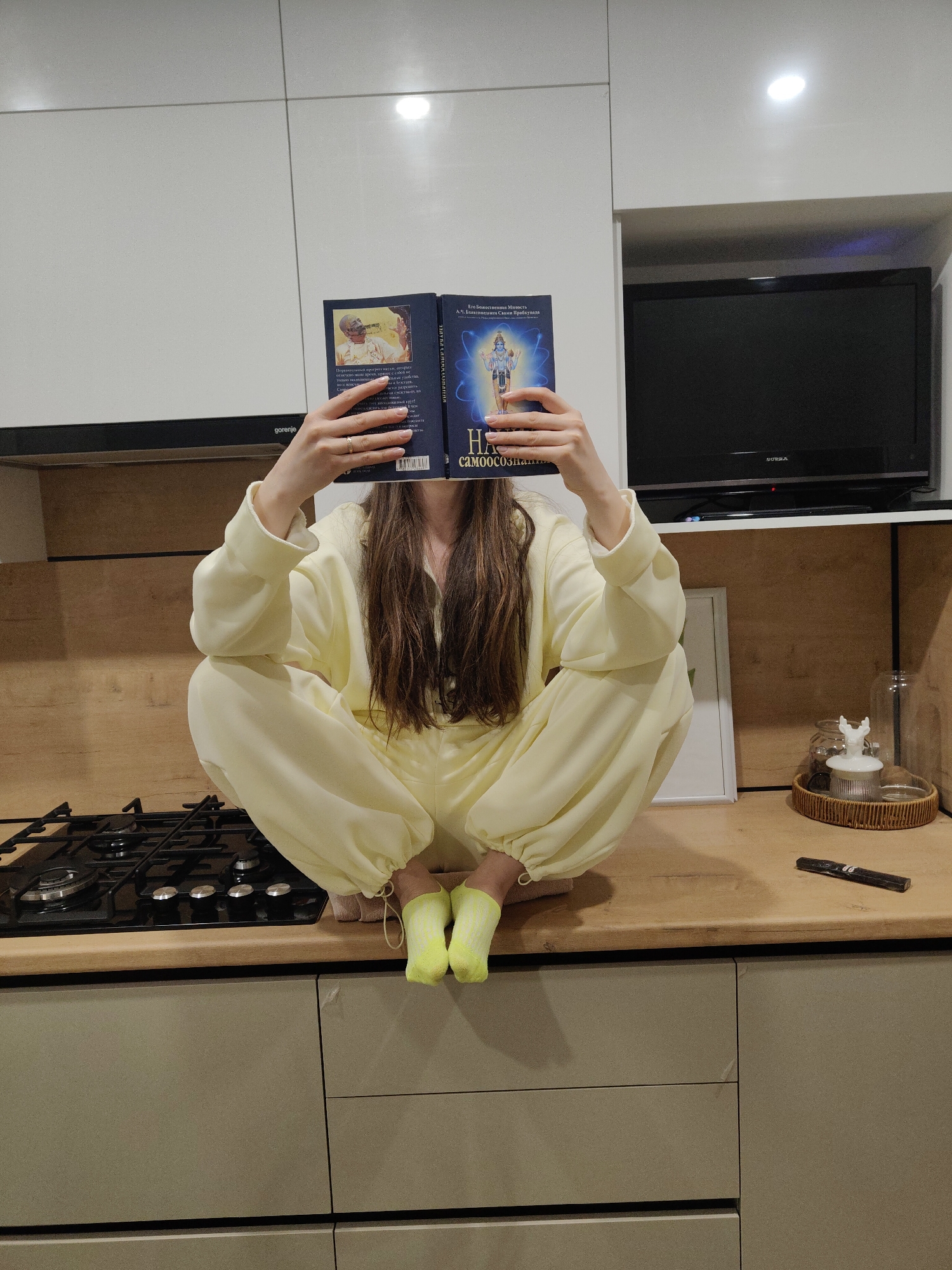 A person is depicted in a modern kitchen, sitting in a crouched or squatting position on a wooden countertop. The individual, whose face is entirely obscured by an open book they are holding, is dressed in pale yellow loungewear or pajamas with elasticated cuffs at the wrists and ankles. They are wearing bright, almost neon, yellow or green socks. Long, dark brown hair cascades down from behind the book.
The book is open, with the right page featuring the title "НА САМООСОЗН..." (likely "На Самоосознание" or "On Self-Consciousness"). Above the title, text reads "Его Божественная Милость А.Ч. Бхактиведанта Свами Прабхупада," indicating the author. An illustration of a blue-skinned deity, possibly Krishna or Vishnu, surrounded by light and cosmic imagery, is prominent on this page. The left page shows smaller text and a black and white portrait of a man, presumably the author. A barcode is also visible on the left page.
The kitchen features white upper cabinets, a black Gorenje extractor hood, and a built-in gas hob with black burners and silver knobs. To the right of the person, a small black flat-screen television with "AOTRA" visible on its frame is placed in an open shelf unit above the countertop. Further right on the countertop, a wicker tray holds glass jars, one topped with a white deer figurine, and a dark, cylindrical object that could be a remote control or pen. The lower cabinets are a light gray color. Artificial lighting, including a ceiling spotlight, illuminates the scene, suggesting it might be evening or night.