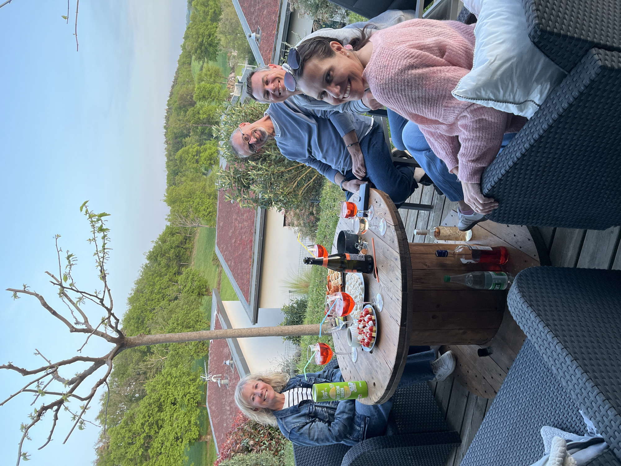 The image captures a social gathering on a wooden deck, likely a patio, in Chambœuf, France, based on the presence of typical French architectural styles in the background. Five individuals are visible, engaged in a relaxed outdoor setting. A blonde woman in a denim jacket sits at a large, circular wooden table, holding a green can of what appears to be Pringles chips. On the table, there are several glasses of orange-colored Aperol spritz, a bottle of sparkling wine, snacks (likely crackers and tomato skewers), and a silver ice bucket. Two men and one woman are visible behind the table. The first man is wearing glasses and a light blue top, next to him is a man wearing sun glasses and a gray top, and finally there is a woman wearing a pink sweater. The background includes a mix of residential buildings with terracotta roofs and a lush green hillside, suggesting a rural or suburban location on the outskirts of Chambœuf. The sky is a pale, early evening hue. A young tree is rooted next to the table. Wicker outdoor furniture complements the relaxed, convivial atmosphere.