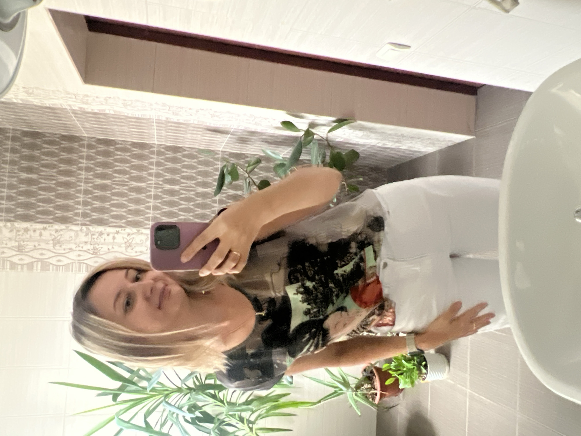 A woman with blonde hair takes a mirror selfie in a bathroom. She is wearing a t-shirt with a graphic print and white jeans. A smartphone in a purple case is held in her right hand. In the background, there are two potted plants, tiled walls, and a doorway. There is no indication of the location being Volgograd, Russia.