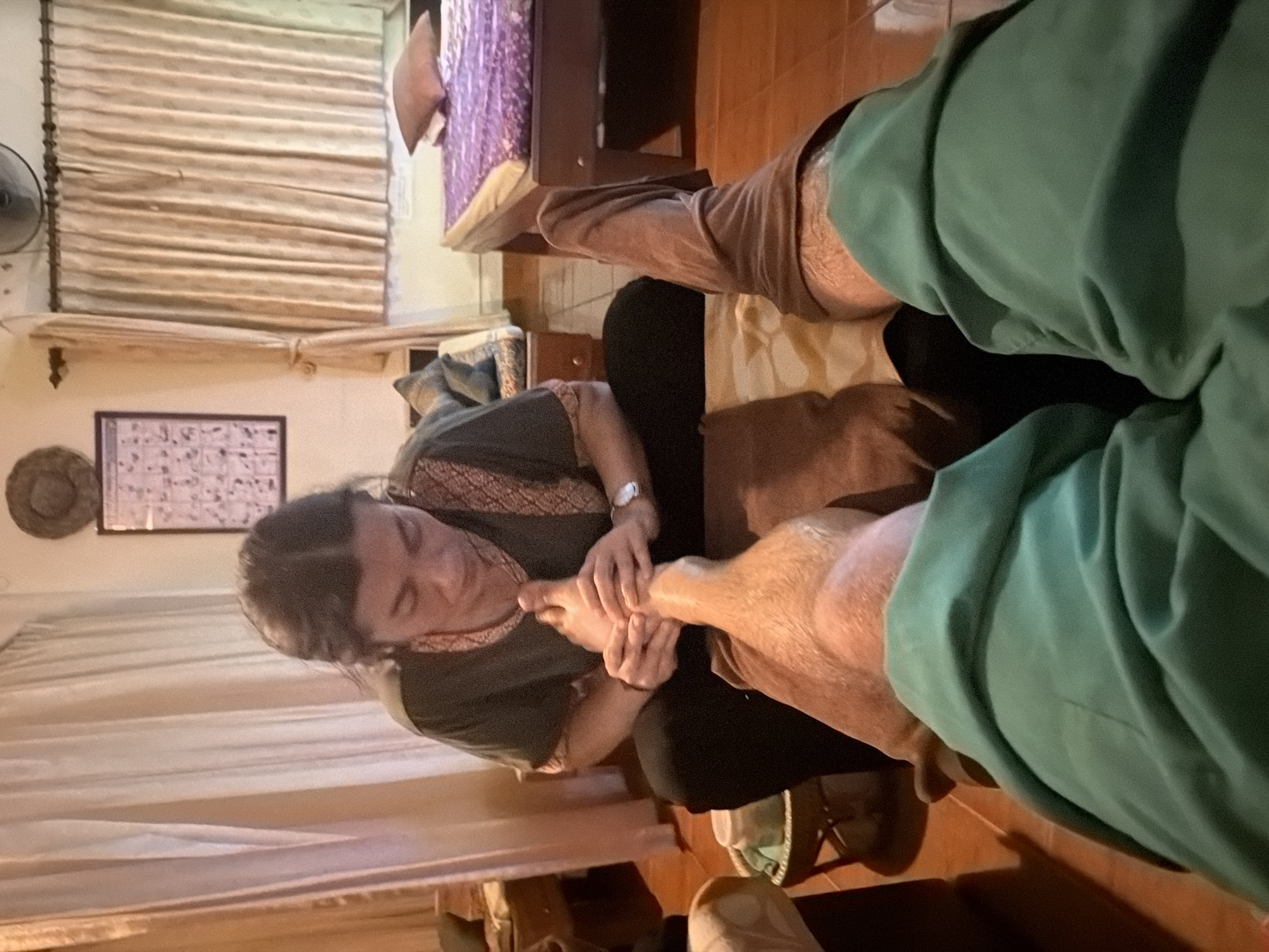 A person is receiving a foot massage in a spa located in Baan Tai, Thailand. The person receiving the massage is lying down, with their legs extended towards the camera, covered by green and brown fabric. The masseuse, wearing a dark green shirt with an ornate neckline, is seated and actively massaging the client's foot with both hands. In the background, there are beige curtains, a framed chart depicting anatomical figures, and a bed with purple linens. A small wooden stool with a container is visible to the left of the masseuse. The floor appears to be tiled. The lighting is warm and ambient, suggesting an indoor setting.