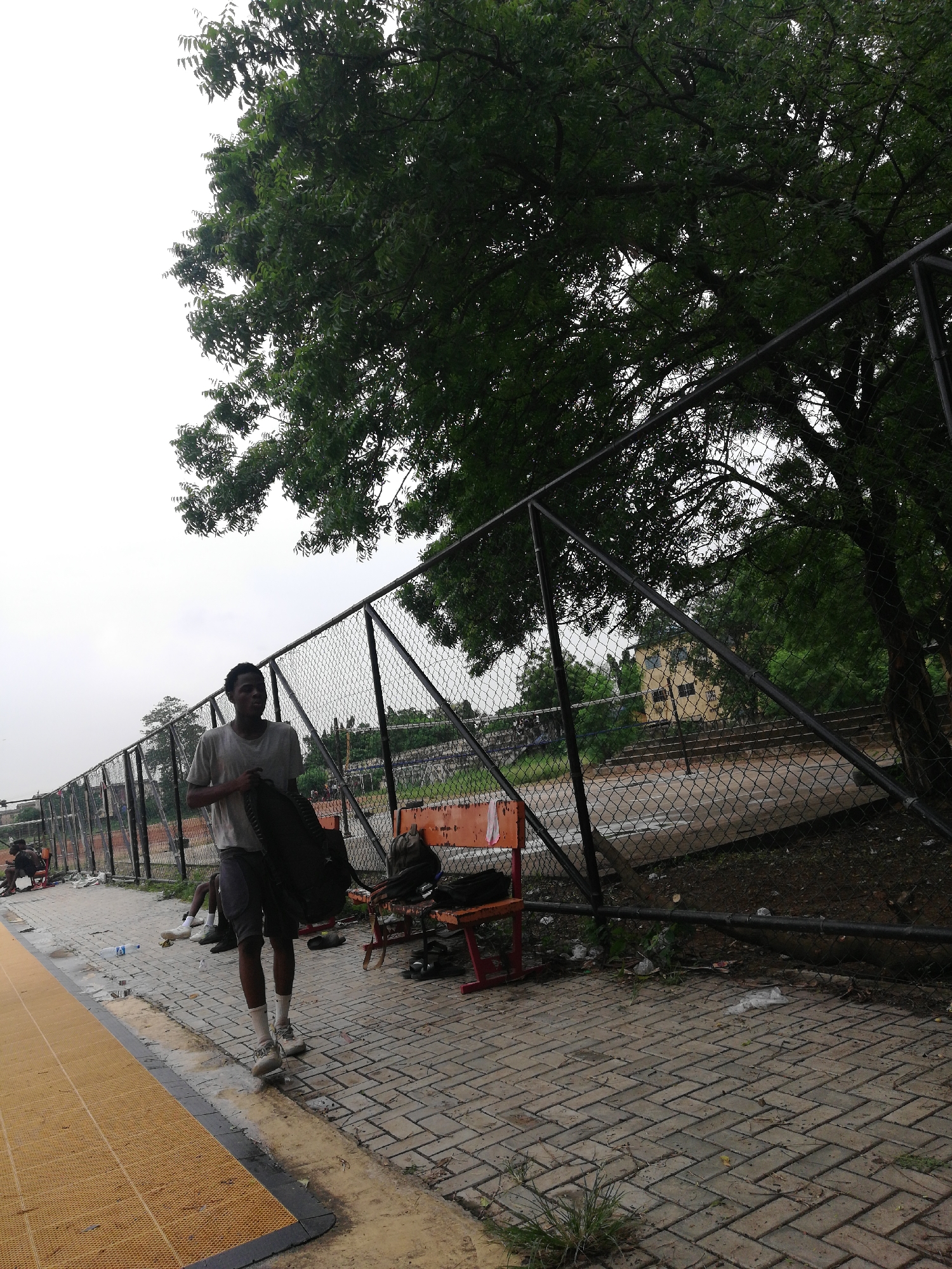 A man carries a backpack on a paved walkway in Lagos, Nigeria. He wears a t-shirt, shorts, white socks, and sneakers. Behind him, a chain-link fence borders a grassy area and a building in the background. Other people are sitting or resting on the walkway and benches near the fence. A large tree hangs over the fence on the right. The sky is overcast.