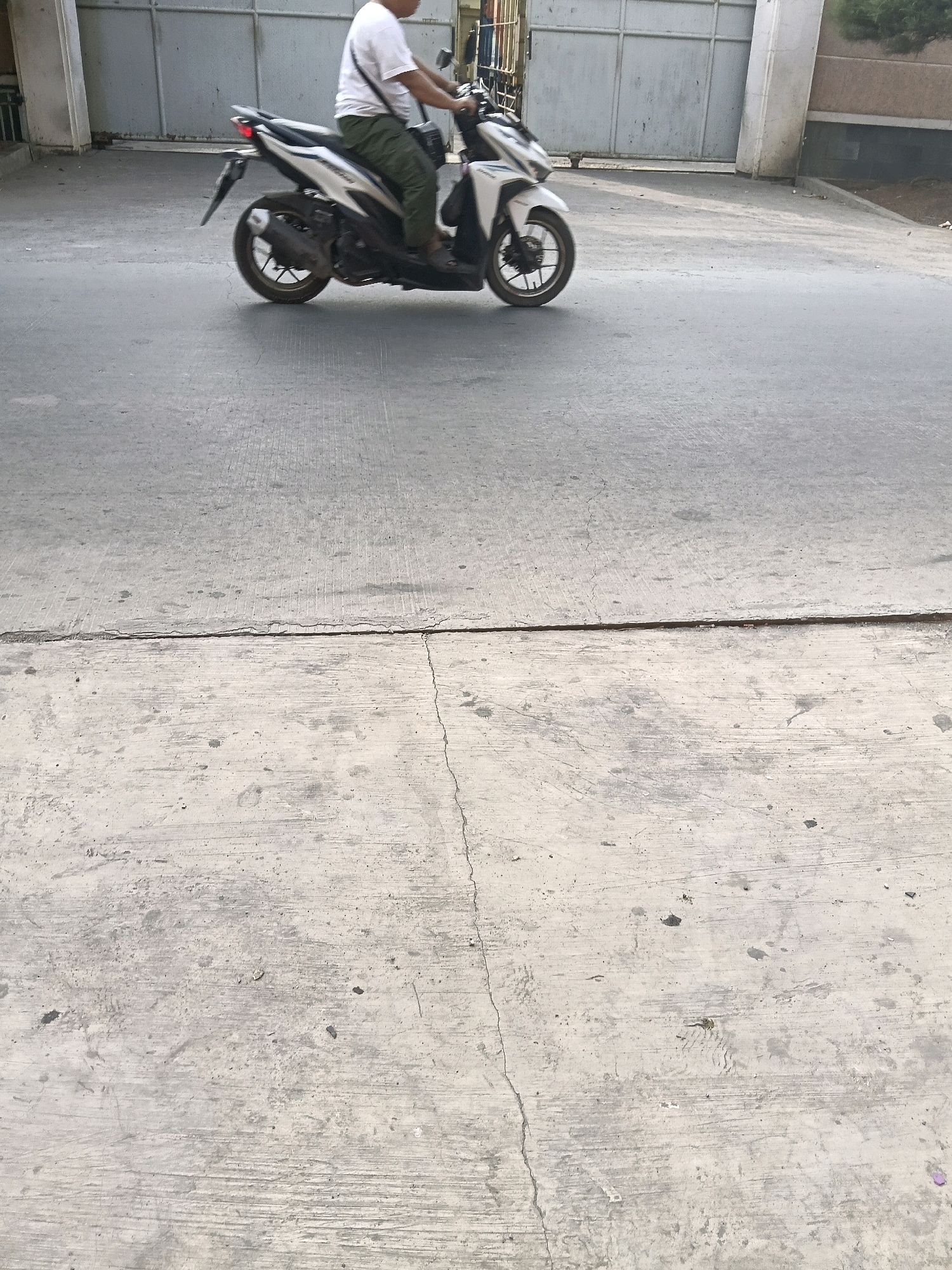 A person is riding a white and silver scooter down a paved road. The rider is wearing a white t-shirt and olive green pants. The scooter has a black seat and black wheels. In the background, there is a large, grey metal fence and a building with a beige facade. The road surface appears to be asphalt with visible cracks and patches. The environment suggests an urban or semi-urban setting in Belimbing, Indonesia.