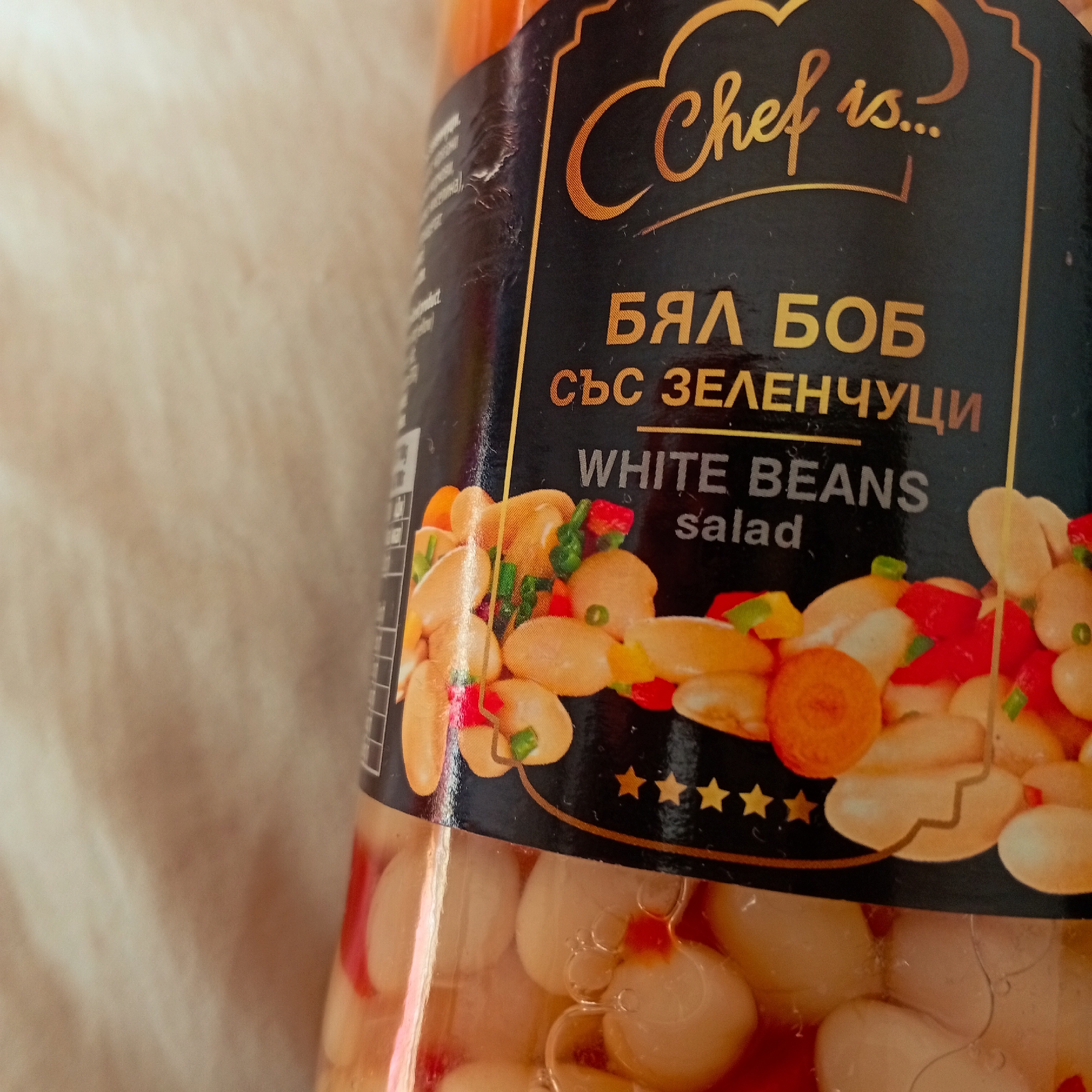 The image displays a close-up view of a clear glass jar containing a food product, positioned on an indistinct light-beige surface in Harmanli, Bulgaria. The jar features a black label with gold and white text. The brand name "Chef is..." is visible in gold script at the top. Below this, the Bulgarian text "БЯЛ БОБ със ЗЕЛЕНЧУЦИ" is printed in gold, with the English translation "WHITE BEANS salad" directly underneath in white. An illustration on the label depicts white beans mixed with chopped red peppers, green fragments, and orange carrot slices. Five gold stars are visible below this graphic. The label shows minor creasing or damage on its left edge. Through the transparent glass, numerous white beans and scattered pieces of red vegetable are discernible, suspended in a clear, reddish liquid. Several small air bubbles are present within the jar's contents. No individuals or specific actions are depicted.