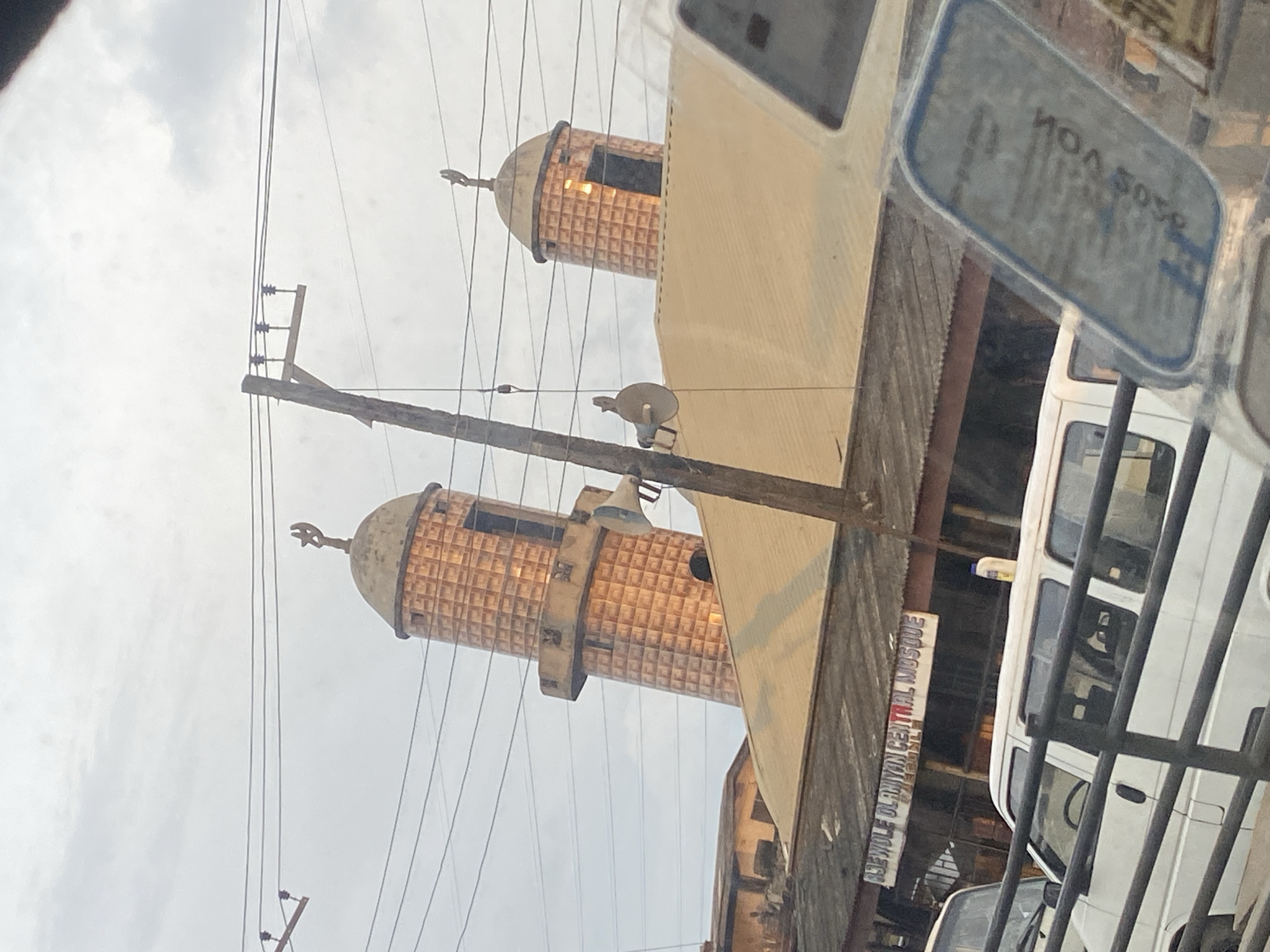 The image shows a view of the "ASEWOLE OLANIYAN CENTRAL MOSQUE" in Ikeja, Nigeria. The mosque features two distinctive cylindrical minarets with a conical roof and a crescent moon finial on top of each. The minarets are tiled with a warm, coppery-brown material that reflects the ambient light. Loudspeakers are mounted on poles near the minarets, suggesting their use for the call to prayer or announcements.
Below the minarets, a tan-colored roof covers the main structure of the mosque. A portion of a sign that reads "ASEWOLE OLANIYAN CENTRAL MOSQUE" is visible on the building's facade. In the foreground, the top of a white van is visible, partially obscuring the view of the mosque's lower levels. The van appears to be parked on a street, with a black metal railing in front of it. Overhead, power lines crisscross the sky, supported by wooden utility poles, indicating an urban setting. The sky is overcast, with diffused light. The overall scene suggests a typical street view in a Nigerian city, with religious architecture and everyday infrastructure coexisting.