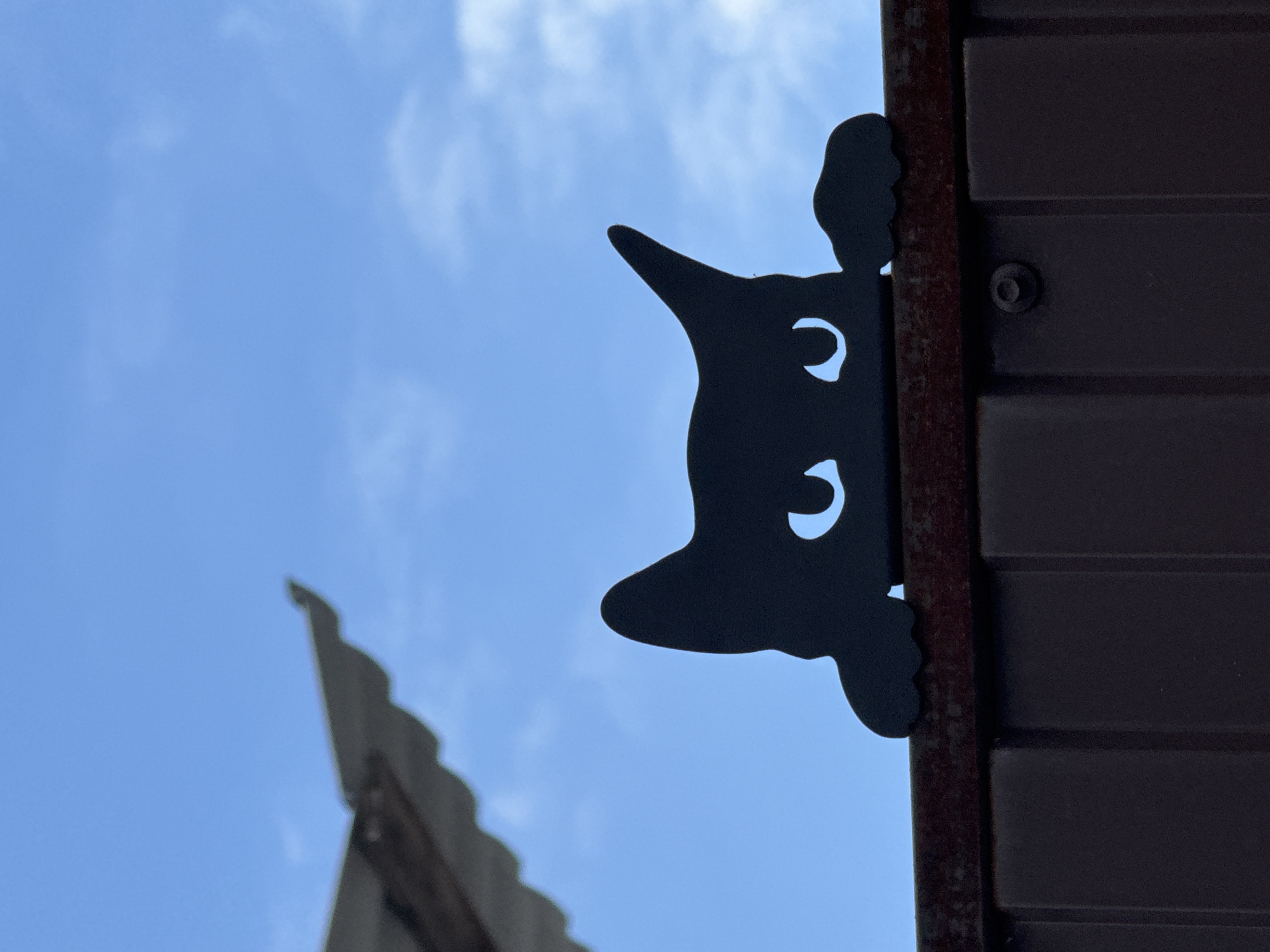 A low-angle shot shows a dark-colored metal gate or door with horizontal grooves, possibly a garage door. Attached to the side of the gate, there is a metal silhouette of a cat peering over the edge, with two moon-shaped cutouts for eyes. In the background, there is a blue sky with a slightly blurred roofline visible to the left. The location is unknown, but the city specified is Steklyannyi, Russia. No people or interactions are visible. The focus is on the gate detail and the cat silhouette.
