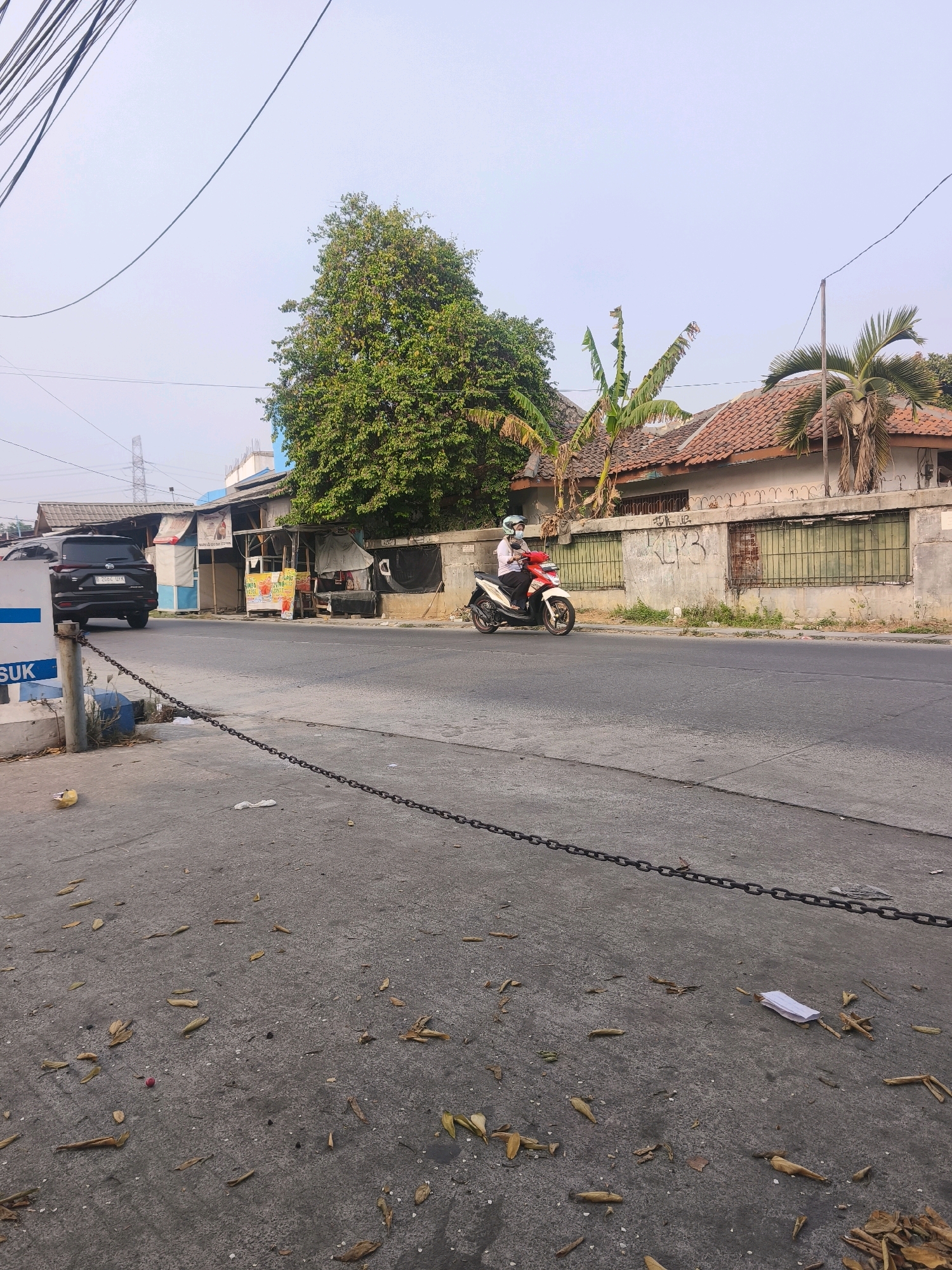 The image depicts a street scene in Bekasi, Indonesia. A black SUV is parked on the left side of the road. Further down the road, a person is riding a red and white scooter, wearing a helmet and a face mask. Several small buildings with tiled roofs line the right side of the road, with some vegetation visible. The road surface is asphalt, and the sidewalk is concrete. The scene is observed from a low angle, close to the ground, with fallen leaves scattered on the sidewalk. A metal chain barrier runs along the edge of the sidewalk. The sky is overcast.