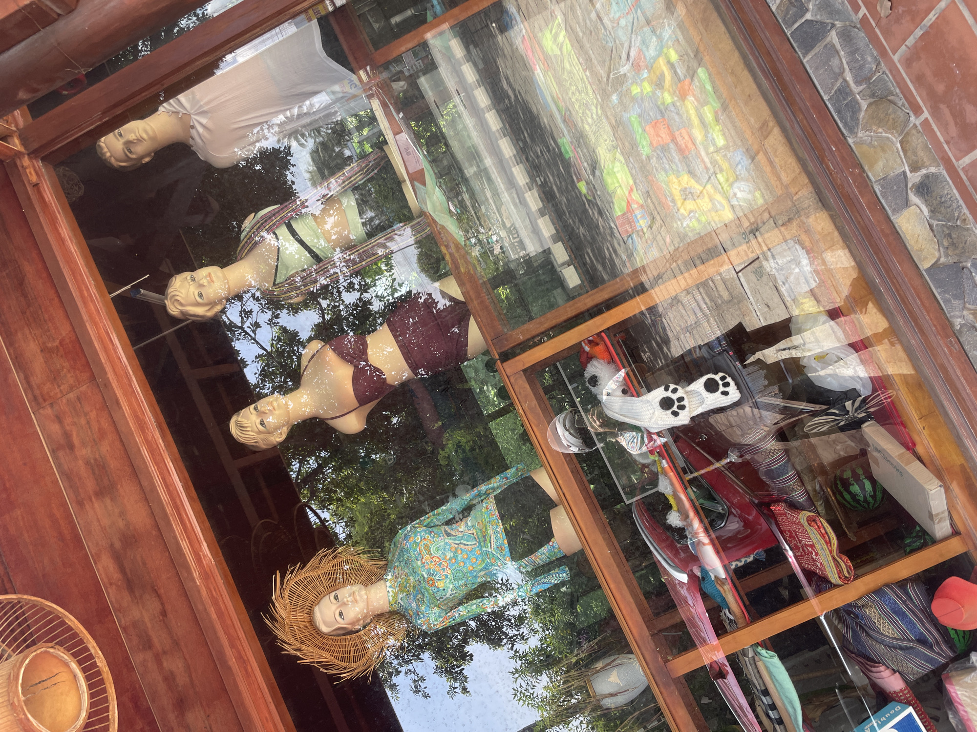 The image shows a storefront in Mũi Né, Vietnam, with mannequins displaying swimwear and clothing. The storefront has a wooden frame and glass windows. Inside, several mannequins are visible. On the left, a mannequin wears a colorful long-sleeved rash guard and black shorts, topped with a wide-brimmed straw hat. Next to it, a mannequin is dressed in a maroon bikini. Further back, another mannequin wears a green and black striped top with a colorful scarf. The fourth mannequin, on the far right, is dressed in a white v-neck shirt. Below the mannequins, shelves are stocked with various items, including textiles with patterned designs, hats, and what appear to be beach towels or sarongs. Some of the items on display include a red car-shaped object, and a white knitted item with paw prints. The reflection on the glass indicates an outdoor setting with trees and foliage visible in the background, suggesting the store is located near natural surroundings. The flooring outside the store is made of stone tiles and bricks.