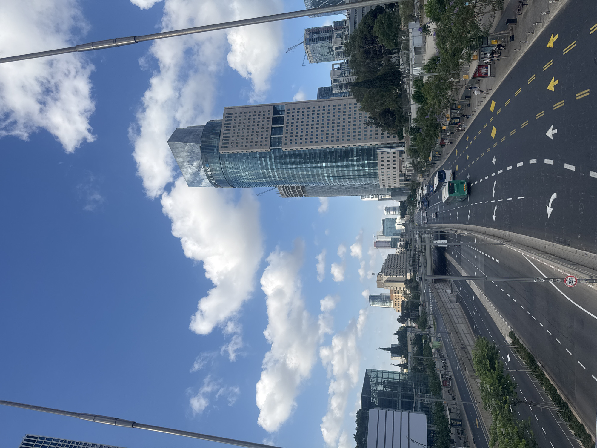 The image captures a wide, multi-lane highway running through a modern urban landscape. The highway is bordered by concrete barriers and trees. Several cars and buses are visible on the road. In the background, a cluster of contemporary skyscrapers dominates the skyline, with construction cranes indicating ongoing development. The sky is a clear blue with scattered white clouds. On the left side of the highway, there are bus stops with people waiting. The overall scene suggests a busy metropolitan area in Tel-Aviv, Israel, possibly depicting the Ayalon Highway.