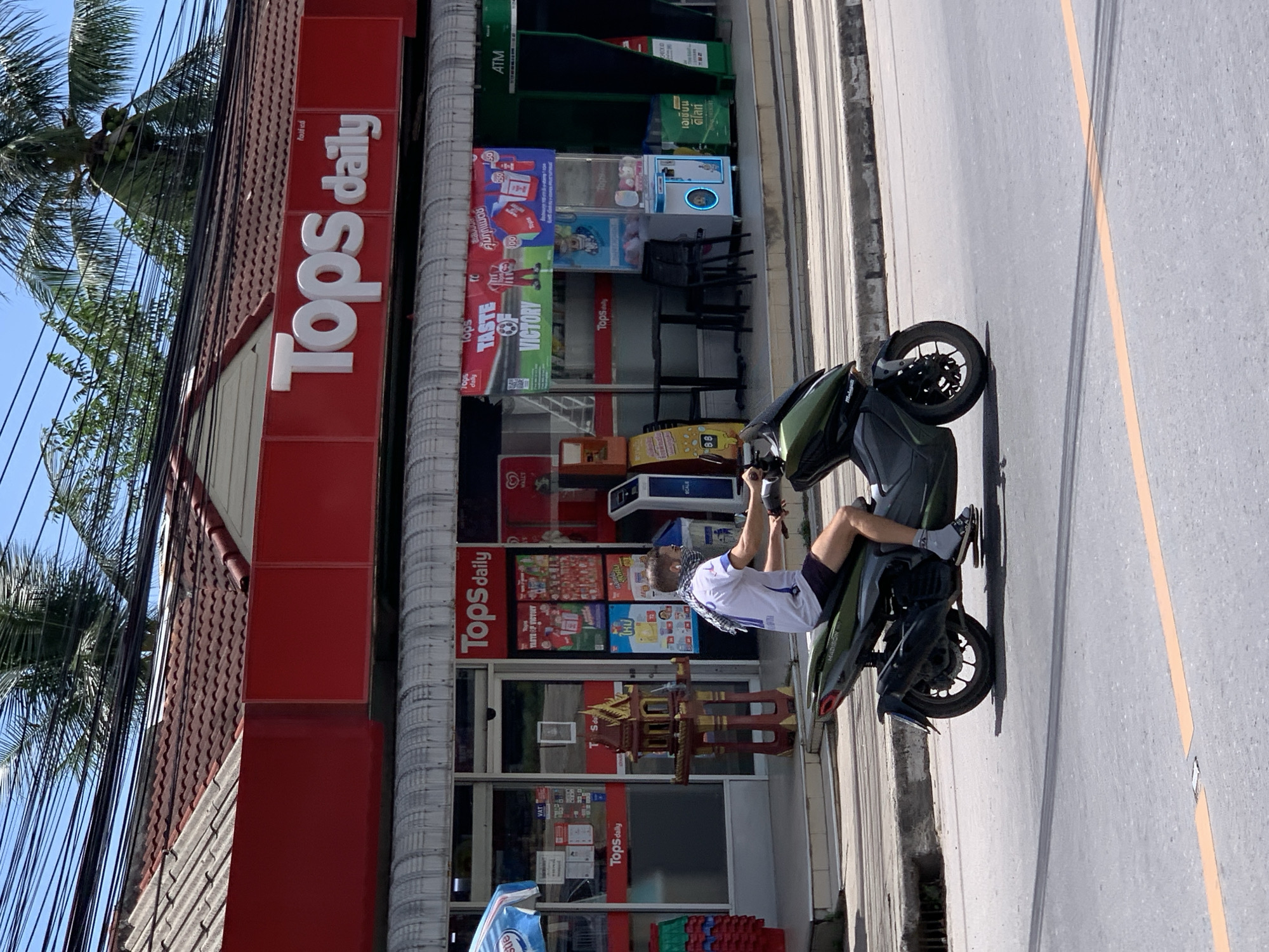 A man wearing a face mask and a white shirt with dark shorts rides a dark green scooter on a paved road. He is in motion, riding towards the right side of the frame. Behind him, a building with a red facade and the "Tops daily" logo is visible. The building appears to be a convenience store or supermarket. There are palm trees and overhead wires in the background. The scene is outdoors, under a clear blue sky, suggesting a warm climate. The street is lined with a sidewalk and a concrete barrier. A small, ornate shrine sits on the sidewalk in front of the store. Several vending machines are also present outside the store. The road has a painted orange line down the center.