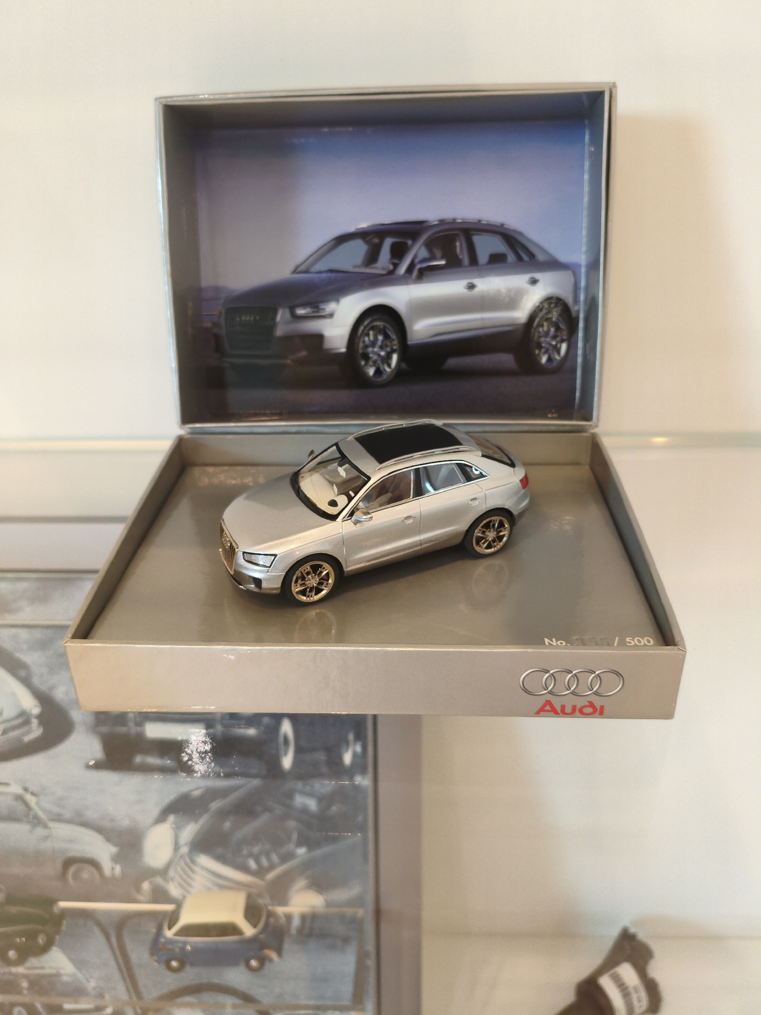 The image shows a display of Audi model cars. In the foreground, a silver Audi Q2 model is placed inside a grey box with the Audi logo and "No. 250 / 500" printed on it. Behind the Q2 model, a larger image of a silver Audi car is displayed within a shadow box. The background features a blurred montage of vintage car images. The overall scene suggests a retail or collector's display, likely located in Ingolstadt, Germany, given Audi's headquarters are there. There are no people or actions depicted in the image.