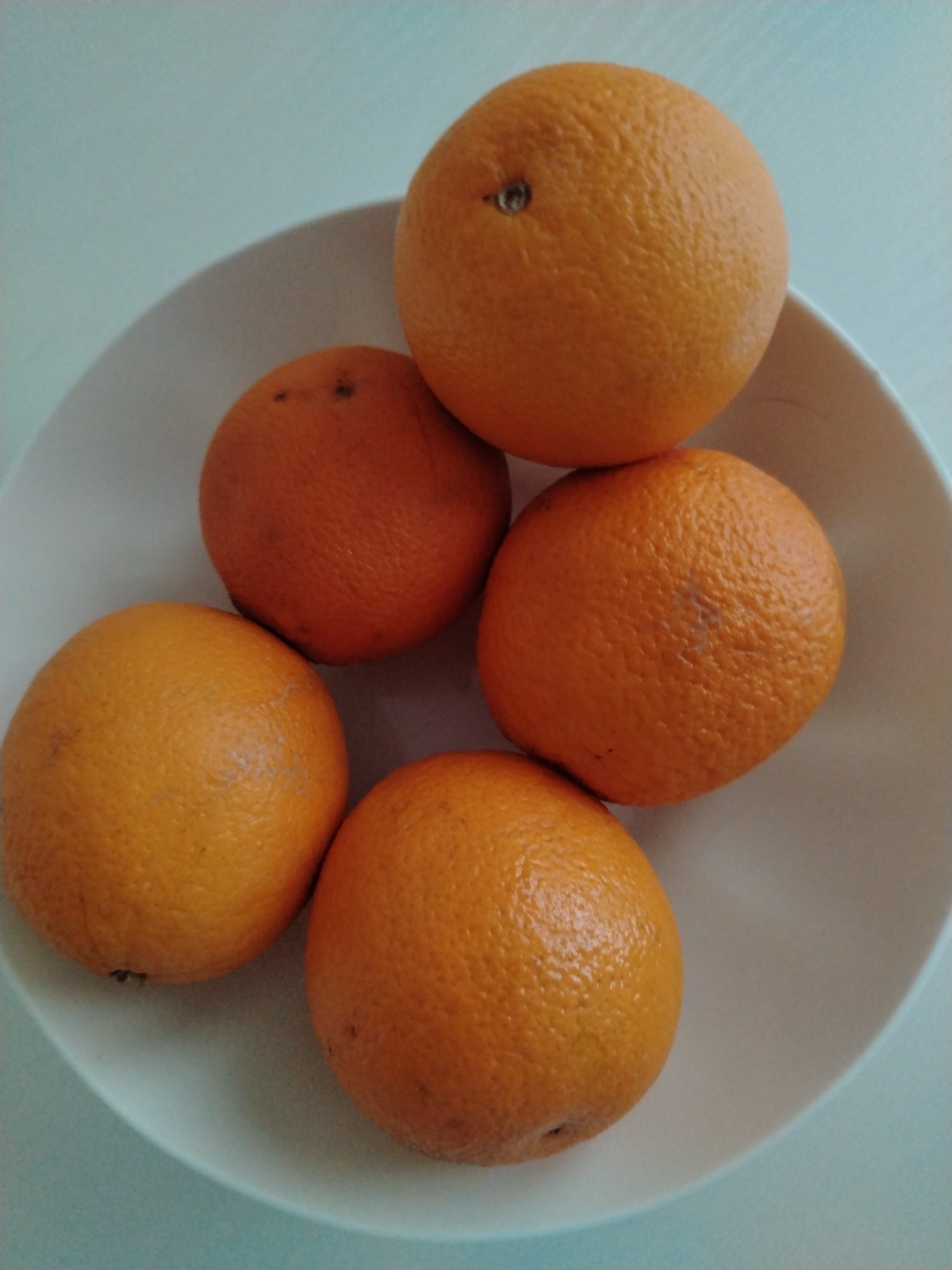 The media file displays a top-down view of a white bowl containing five oranges. The oranges are a vibrant orange color and have a textured peel. One orange is positioned at the top of the bowl, slightly off-center to the right. Below it, and slightly to the left, are two more oranges. The bottom of the bowl contains two more oranges, one centrally located and the other to its left. The bowl itself is white and has a smooth, curved surface. The background is a plain, light blue surface, which appears to be a table or countertop. There is no discernible text in the image. The lighting suggests it may be daytime, possibly indoors due to the consistent illumination and lack of strong shadows. The overall scene is simple and focused on the fruit.