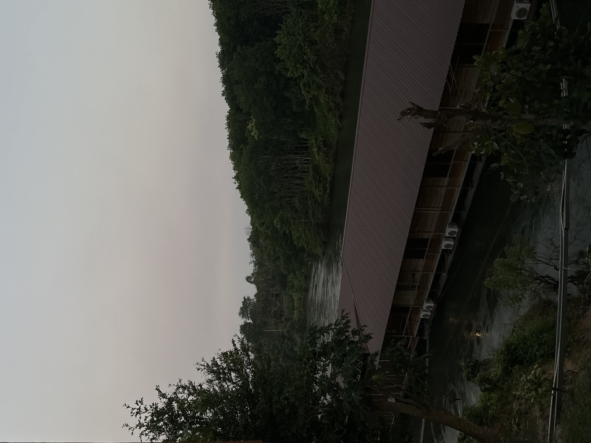 This is an outdoor scene depicting a river running alongside a building with a red roof, possibly a resort or guesthouse in Sai Yok, Thailand. The sky is overcast. Dense green vegetation covers the opposite bank of the river, and trees are visible in the foreground. The building is constructed with wooden beams and has air conditioning units mounted on its exterior. The river water appears slightly turbulent. The scene appears to be taken from ground level.