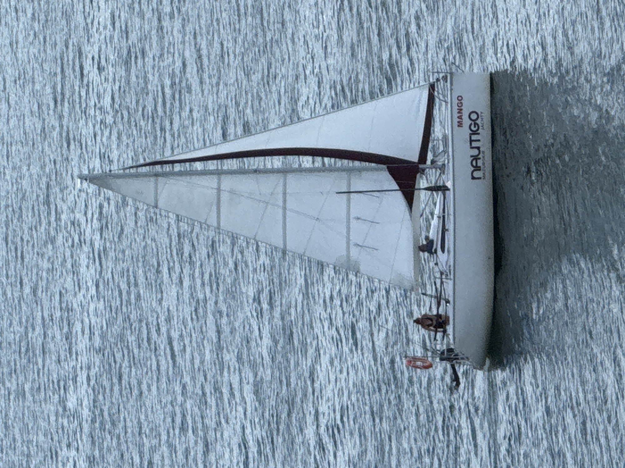 This media file shows a sailboat on a body of water, likely a lake given the location context of Mikołajki, Poland. The sailboat has a white sail with a dark red trim along the leading edge. The hull of the sailboat is also white and appears to have some text printed vertically on its side. The text reads "MANGO" at the top and "NAUTIGO BALTUS HOUSE" below it. The water is choppy, with many small ripples, suggesting a breezy day. The lighting indicates it is daytime, likely with overcast skies or indirect sunlight, as there are no strong shadows visible. A few people are visible on the sailboat, but they are not distinct enough to identify details. The scene depicts a sailing activity, likely recreational. The overall impression is one of a serene, albeit active, day on the water.
