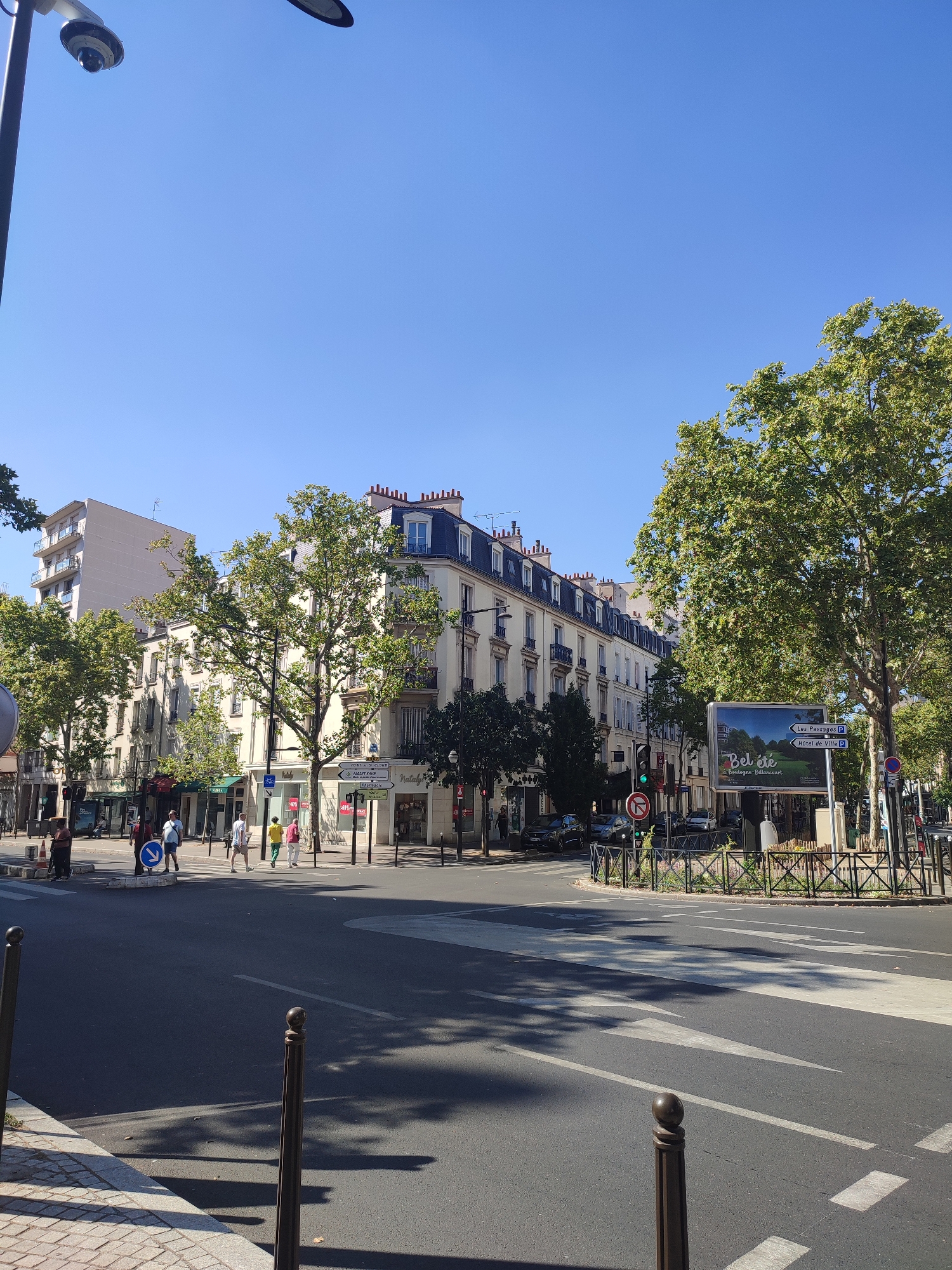 The image shows a street corner in Boulogne-Billancourt, France, on a clear, sunny day. In the foreground, a wide asphalt road features a pedestrian crossing marked with white stripes. Two dark, slender bollards stand on the side of the road, one closer to the camera. In the mid-ground, several people are crossing the street at the designated crosswalk. They are casually dressed, with one person in a blue shirt and shorts, another in a yellow shirt and white pants, and a third in a pink shirt and white pants. To the right of the crosswalk, a circular green space is enclosed by a decorative metal fence. A large billboard with an image of a park and text reading "Bel été à Boulogne-Billancourt" is positioned behind the fence. Across the street, a line of multi-story buildings with French architectural features, including mansard roofs and balconies, are visible. The buildings house various shops on the ground floor, indicated by signage and storefronts. Trees with lush green foliage are planted along the sidewalks, casting shadows on the road. A traffic light is visible on the left side of the intersection, showing a green light. A street lamp with a security camera is mounted on a pole in the upper left corner of the image. The sky is a clear, vibrant blue.