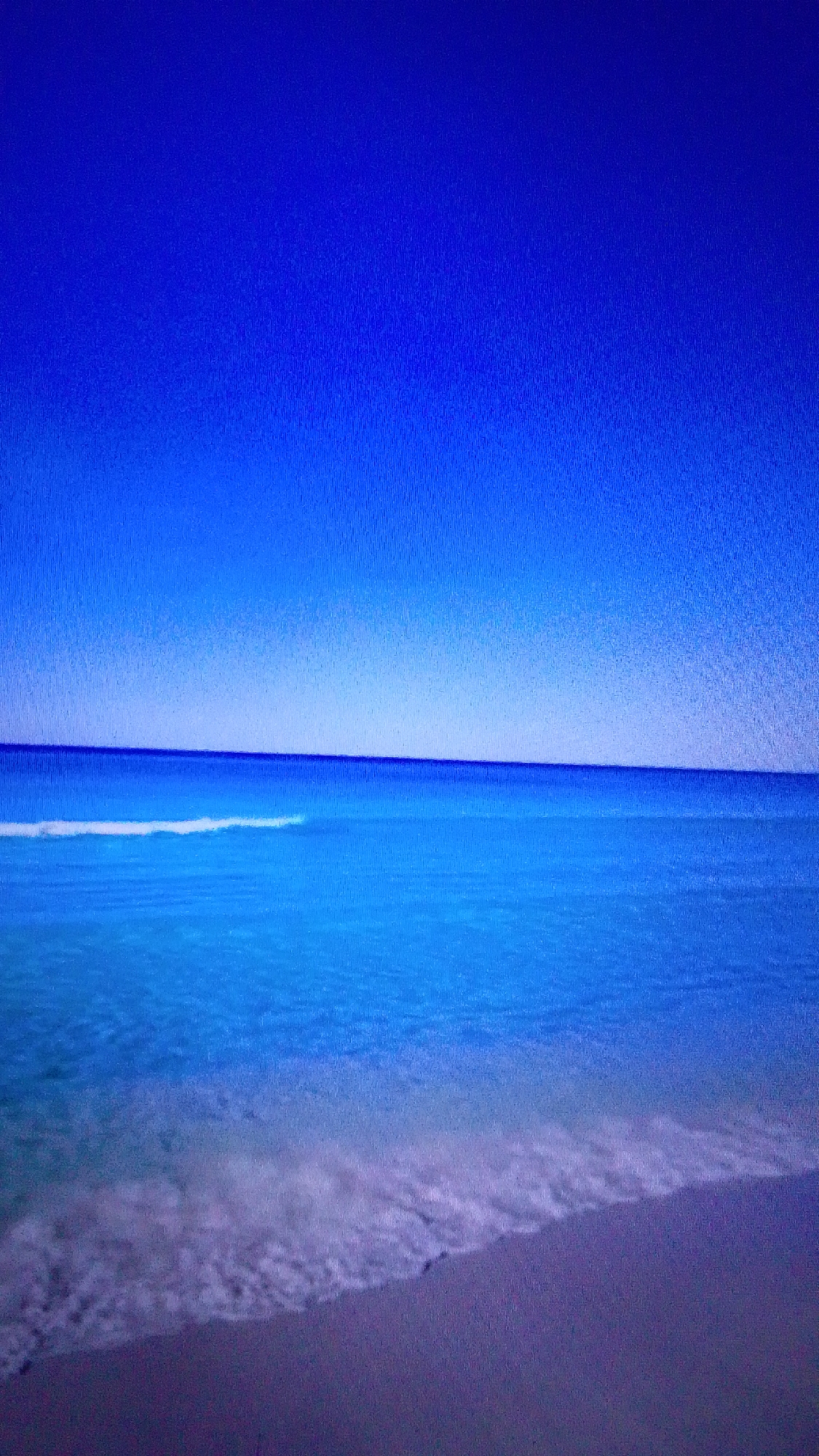 This is an image of a beach with a clear blue sky and calm ocean. The water is a vibrant turquoise, with gentle waves lapping at the shore. The sand is a light brown color, and there are a few scattered shells visible. The image appears to be taken on a sunny day, with the sun casting a warm glow over the scene. There are no people or objects present in the image. The location is not Aznakayevo, Russia, as the image depicts a tropical beach.