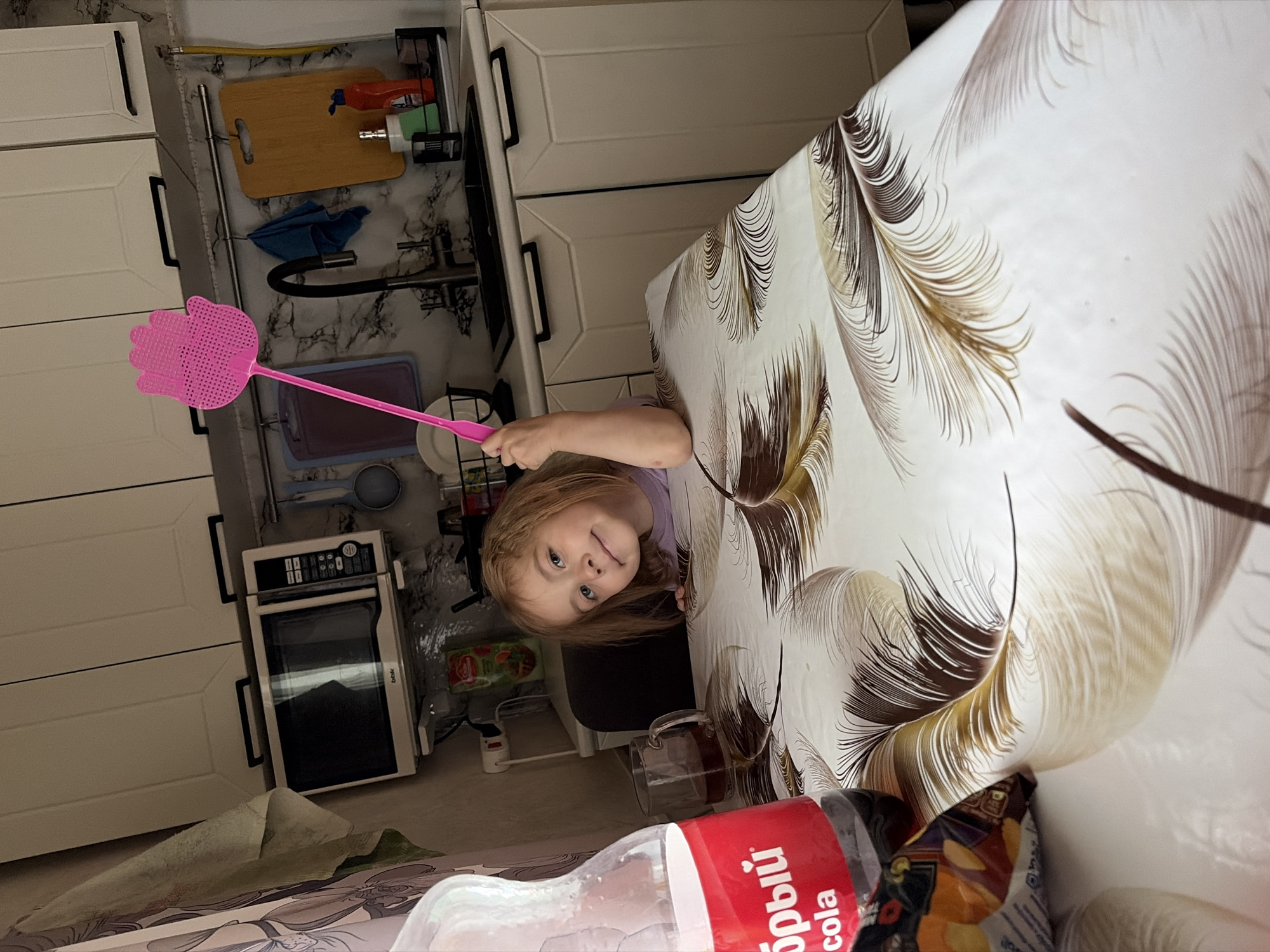 A young girl with blonde hair is sitting at a kitchen table in a home in Tambov, Russia. She is holding a pink fly swatter in her right hand, with her left arm resting on the table. She is looking at the camera and smiling. On the table, there is a bottle of cola, a glass of tea, and a bag of chips. The kitchen cabinets are white, and the countertop is grey. A microwave is mounted above the counter. The scene is brightly lit, suggesting it is daytime.