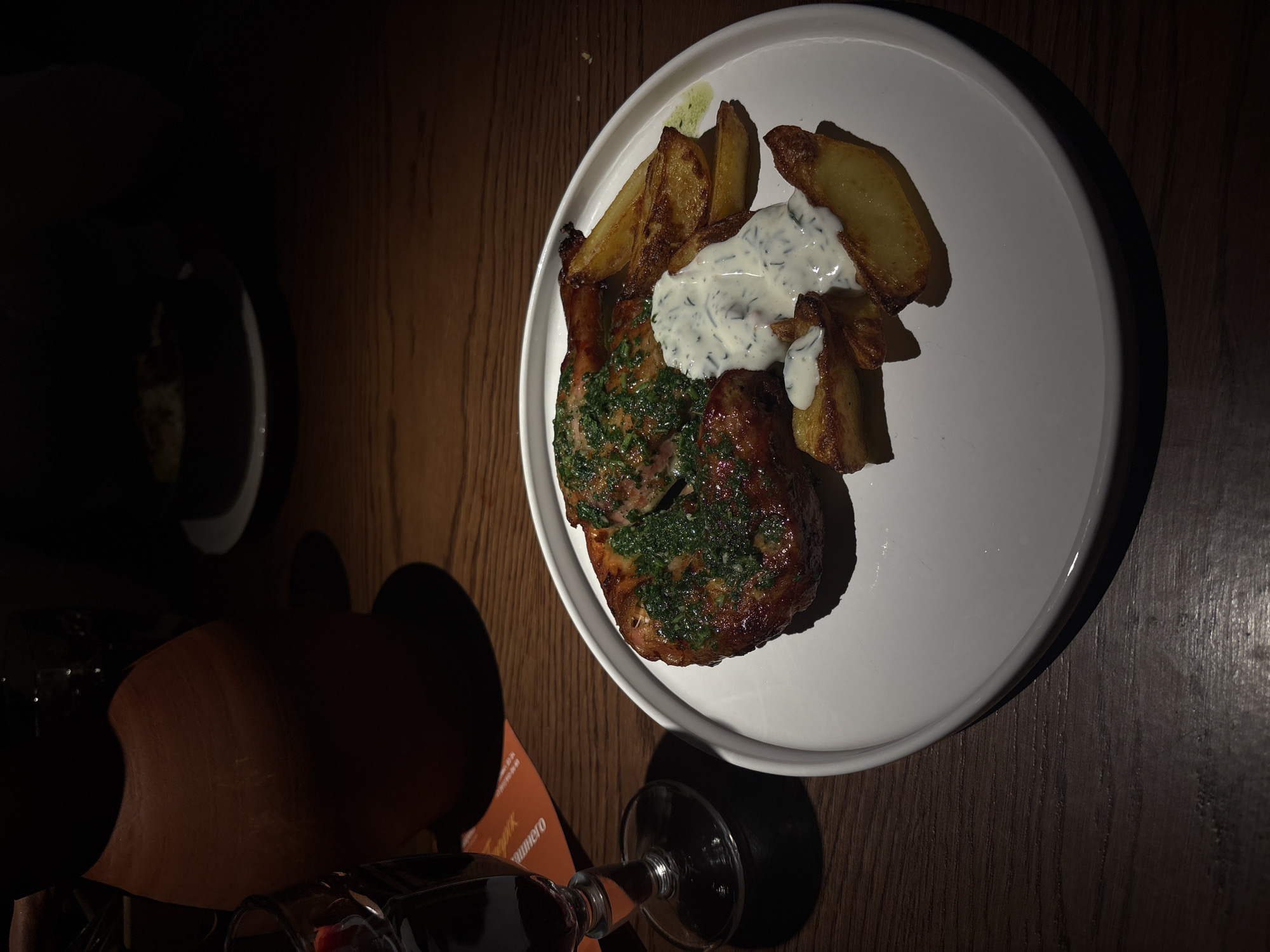 The image captures a plate of food on a dark wooden table, alongside a glass of red wine. The main dish consists of a grilled meat chop, topped with green herbs, and served with potato wedges and a creamy, white sauce. A menu with lettering that appears to be Russian sits nearby, partially obscured by the wine glass. The location is not evident from the image, but it's possible the menu hints at a specific restaurant in Saint Petersburg, Russia. The lighting is dim, creating strong shadows on the table and the adjacent items, including another plate in the background. The scene is likely inside a restaurant or dining establishment.