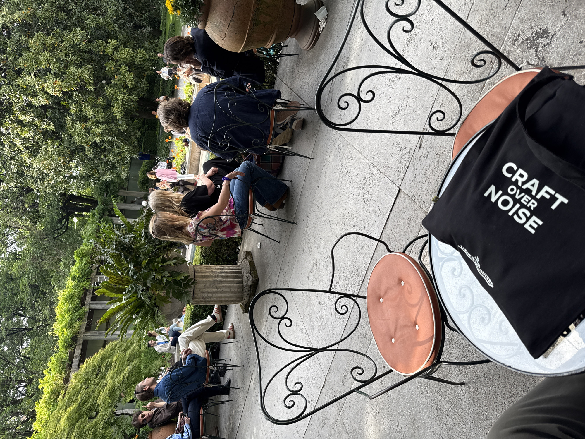 This image shows an outdoor seating area, likely a cafe or restaurant, situated in a garden or park-like setting. There are several people seated at tables, some of whom appear to be engaged in conversation. The furniture consists of ornate metal chairs and tables. A black tote bag with the words "CRAFT OVER NOISE" is visible on one of the tables. The location is presumably Milan, Italy. The scene suggests a relaxed, social gathering in a pleasant outdoor environment.