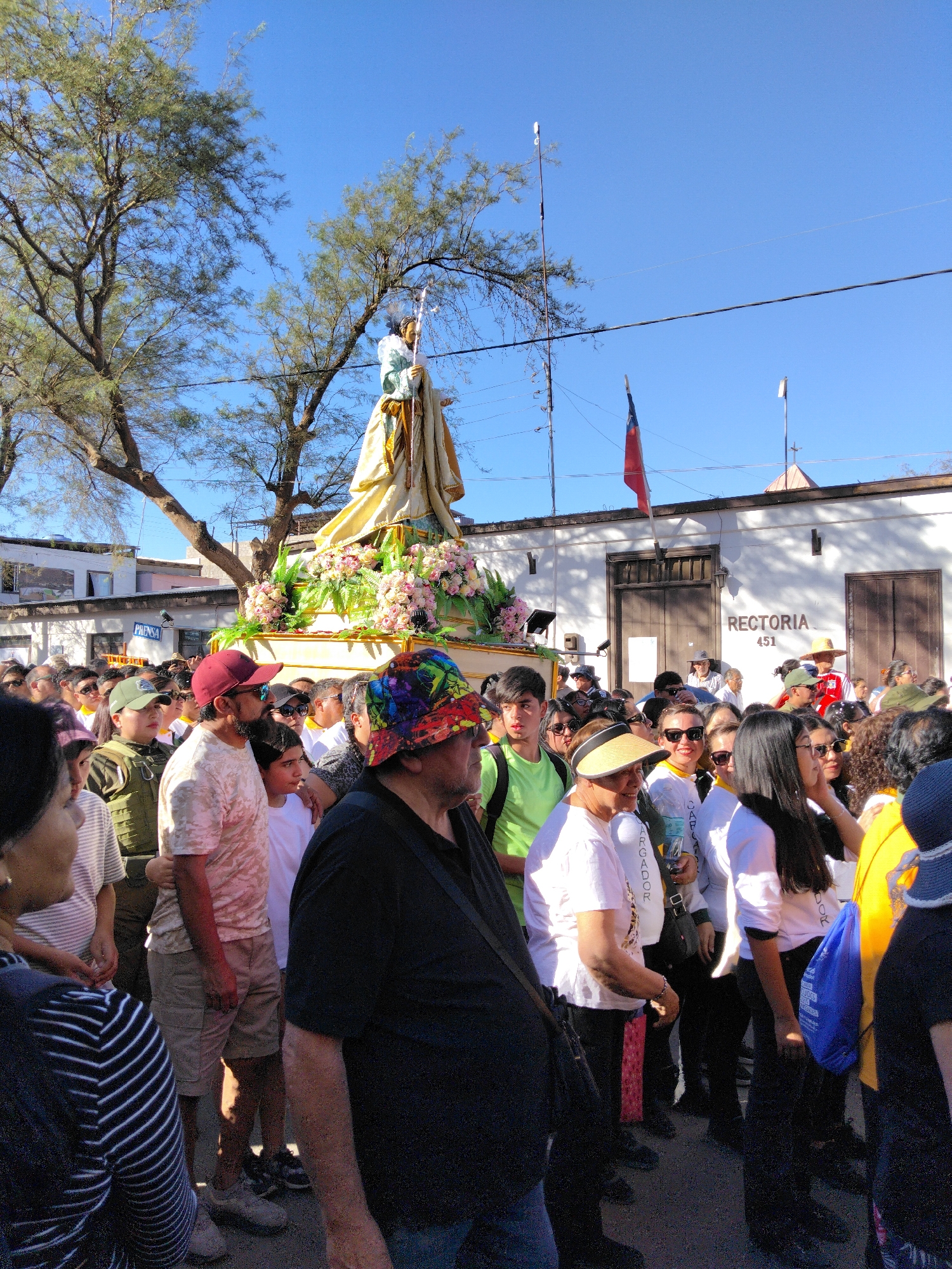 A religious procession is taking place outdoors, likely in the city of Pozo Almonte, Chile, given the context of the "Rectoria 451" sign visible on a building in the background, which is a known address for the main church there. A large, ornate float carrying a statue of a saint, adorned with pink and white flowers, is the central focus, partially obscured by a crowd of people. The statue appears to be dressed in a golden robe and holds a staff.
The procession is moving along a street lined with low-rise buildings. The building with the "Rectoria 451" sign is white with brown wooden doors and architectural details. A Chilean flag is visible on a flagpole in front of this building. Sunlight casts shadows, indicating it is daytime.
The crowd consists of many individuals of varying ages, mostly wearing casual clothing. Some are looking towards the float, while others are interacting with each other. Several people are wearing hats, including a man in the foreground with a brightly colored, patterned hat. A man in a light pink t-shirt and khaki shorts is standing in the foreground, to the left of the man with the colorful hat, and appears to be looking towards the float. Another man in a red baseball cap is to his left. Many people in the crowd are wearing white shirts.
The environment appears to be a public street with trees and power lines overhead. The sky is a clear, bright blue. The overall atmosphere suggests a community event, likely a religious festival or celebration.
