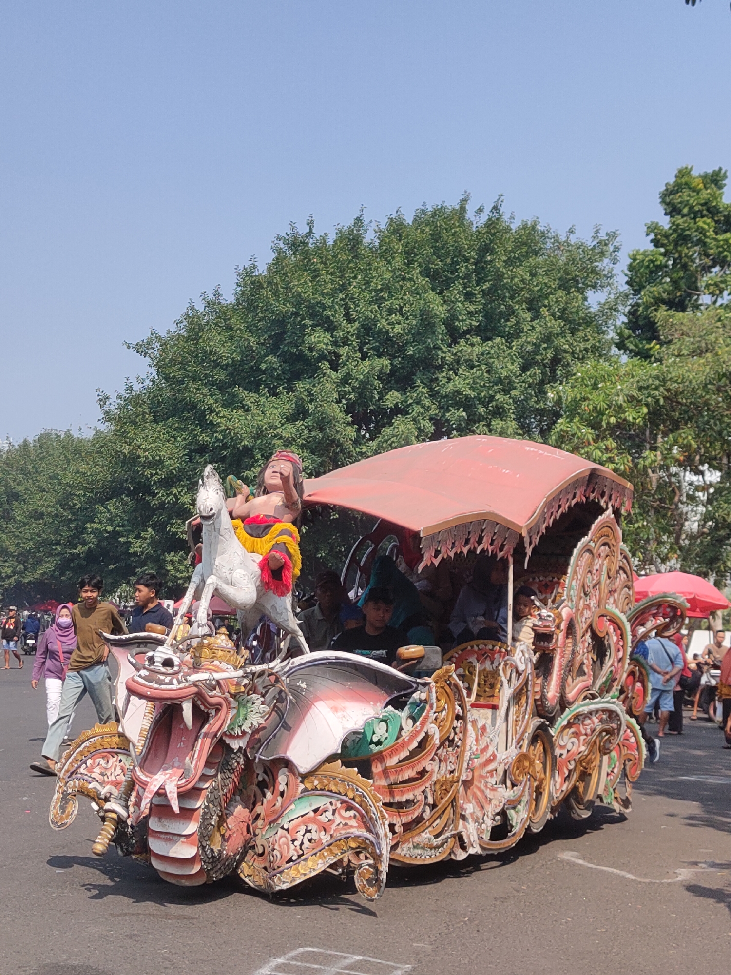 This is a photograph of a colorful, ornate float in motion, likely part of a parade or cultural event, on a paved street in Ngasem, Indonesia. The float is intricately decorated with carved mythical creatures, including a prominent dragon's head at the front with its mouth open and a horse statue with a rider at the rear. The float has a red canopy providing shade for passengers, some of whom are visible seated inside. People are walking alongside and behind the float, indicating a public gathering. The background shows lush green trees and a clear blue sky, suggesting a sunny day. Several individuals, dressed in casual attire, are captured in the frame, some on foot and others near motorcycles. The overall scene depicts a vibrant procession with elaborate cultural artistry and community participation.