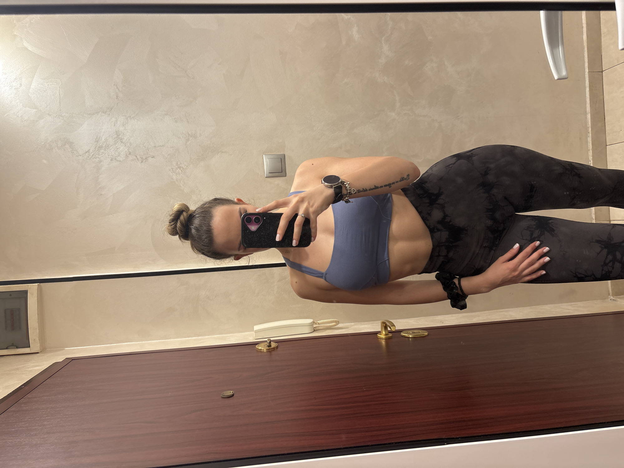 The image displays a mirror selfie of a woman, oriented 90 degrees clockwise. The woman, viewed from her chest to her mid-calf, is captured standing sideways. She wears a blue sports bra and dark, intricately patterned leggings. Her hair is styled in a bun. In her right hand, she holds a smartphone with a black, possibly glittery, case, partially obscuring her face. A smartwatch with a black band is visible on her left wrist, and a ring adorns her left hand. A script tattoo is present on her inner left bicep, and a black scrunchie is on her right wrist.
The background, reflected in the mirror, features an indoor setting. A wall with a light beige or off-white textured finish is visible. On this wall, a silver electrical outlet or switch is present. In the upper right of the reflection, a white, possibly ceramic, object resembling part of a sink or toilet can be seen. Below the wall, a dark reddish-brown wooden surface, likely a countertop or dresser, is reflected. On this surface, a white landline telephone with a coiled cord, a brass-colored door handle or fixture, and a small golden disc are visible. The mirror is framed by a thin black border at the top and bottom. The lighting suggests an indoor environment with artificial light. No explicit text is clearly legible on the tattoo, phone screen, or smartwatch.