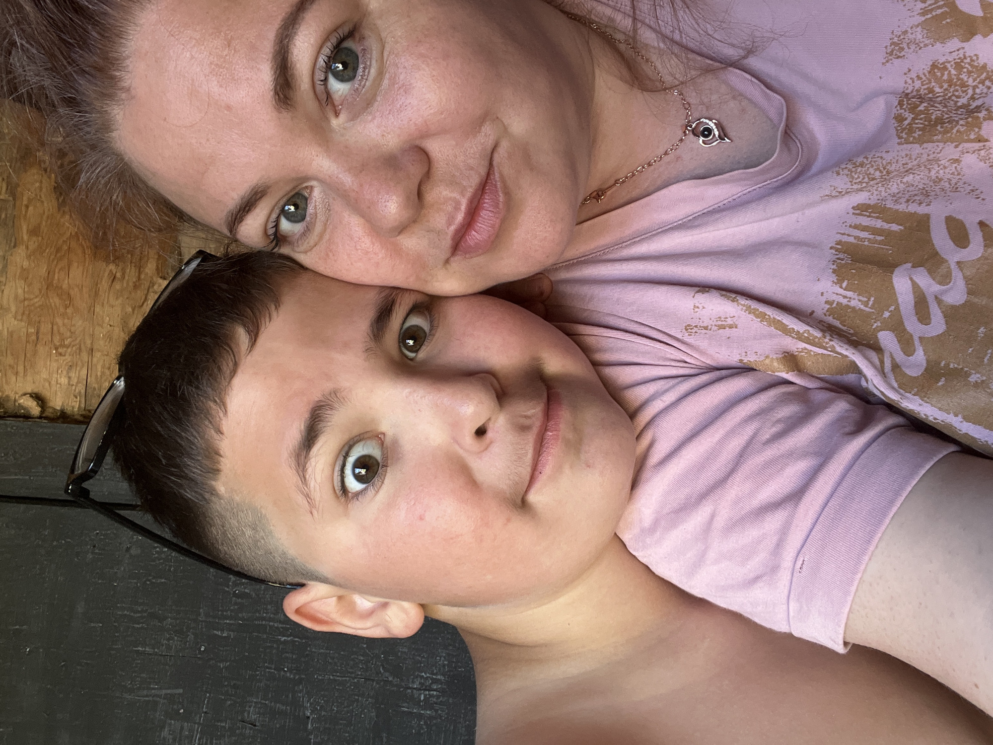 A close-up, selfie-style photograph shows two people, a child and an adult, positioned side-by-side. The child, on the left, leans their head against the adult's shoulder. Both individuals are looking towards the camera, with the child's eyes wide and surprised. The adult, on the right, has a softer expression. The background is indistinct, appearing to be a wooden wall or structure. The child is shirtless, while the adult is wearing a pink t-shirt with a gold graphic design. The adult is also wearing a necklace with a heart-shaped pendant. The overall impression is an intimate, casual moment captured between two individuals. The location is not discernable from the visual information in the image.
