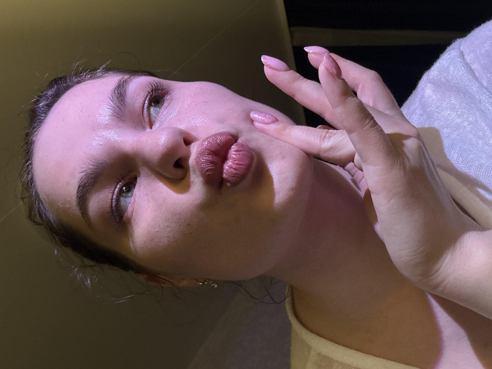 The image shows a close-up of a young woman's face and hand. She has her lips pursed as if to kiss, and her right hand is near her chin, with her fingers extended. Her fingernails are painted a light pink. The lighting in the image is warm and casts a pinkish hue on her skin. Her hair is dark and appears to be pulled back. She is wearing a light-colored garment, possibly a sweater or shirt, with a delicate texture. The background is dark and out of focus, suggesting an indoor setting. There is no discernible city or location information visible in the image.