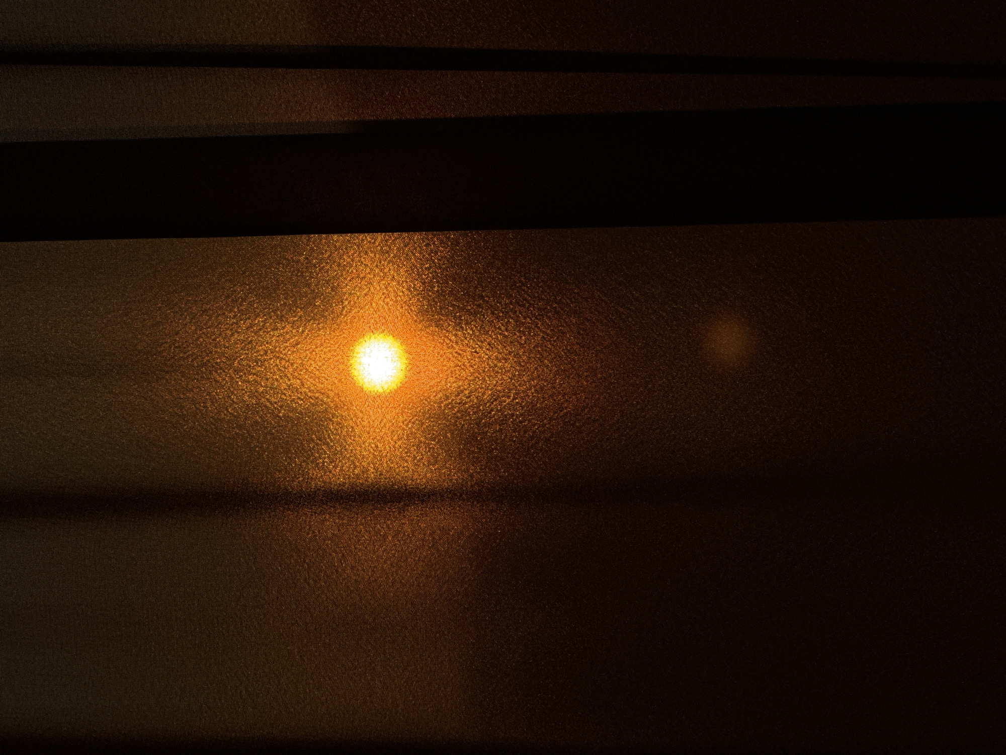 A bright, circular sun is visible through a textured, translucent surface, possibly a frosted glass window or a sheer curtain. The sun is a vibrant yellow-orange, with a halo of diffused light. Dark, vertical elements, likely curtains or blinds, frame the right side of the image, creating deep shadows and obscuring portions of the background. The overall lighting is warm and subdued, suggesting either sunrise or sunset. The surface in front of the sun has a subtle woven texture, contributing to the diffusion of light and creating a speckled effect.