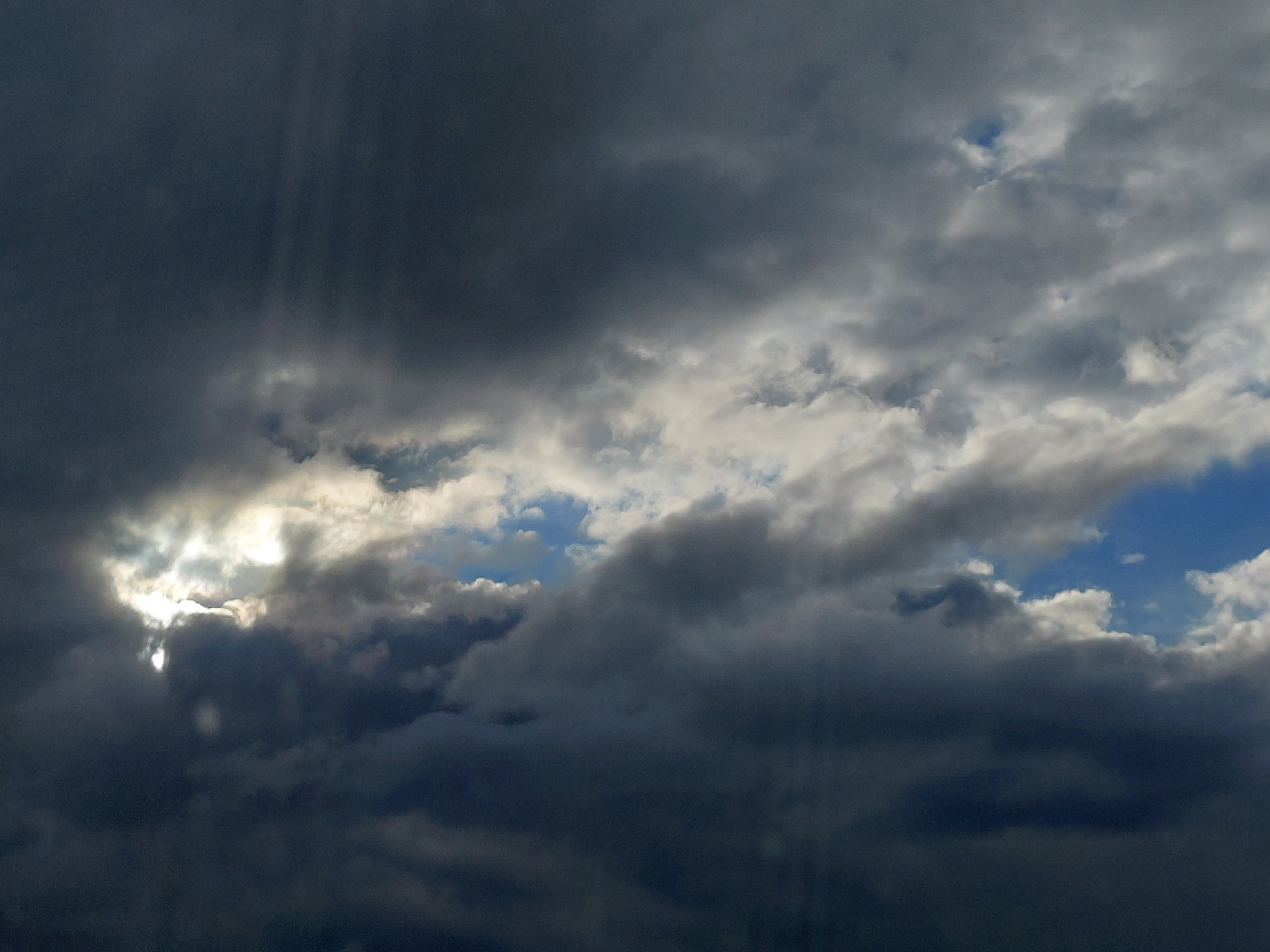 The image depicts a dramatic sky filled with dark, heavy storm clouds. A break in the clouds reveals a patch of bright sunlight, casting rays downwards. The scene is a panoramic view of the sky, with no buildings, people, or any other discernible objects visible. The location in Stromilovo, Russia, is not evident from the image itself.