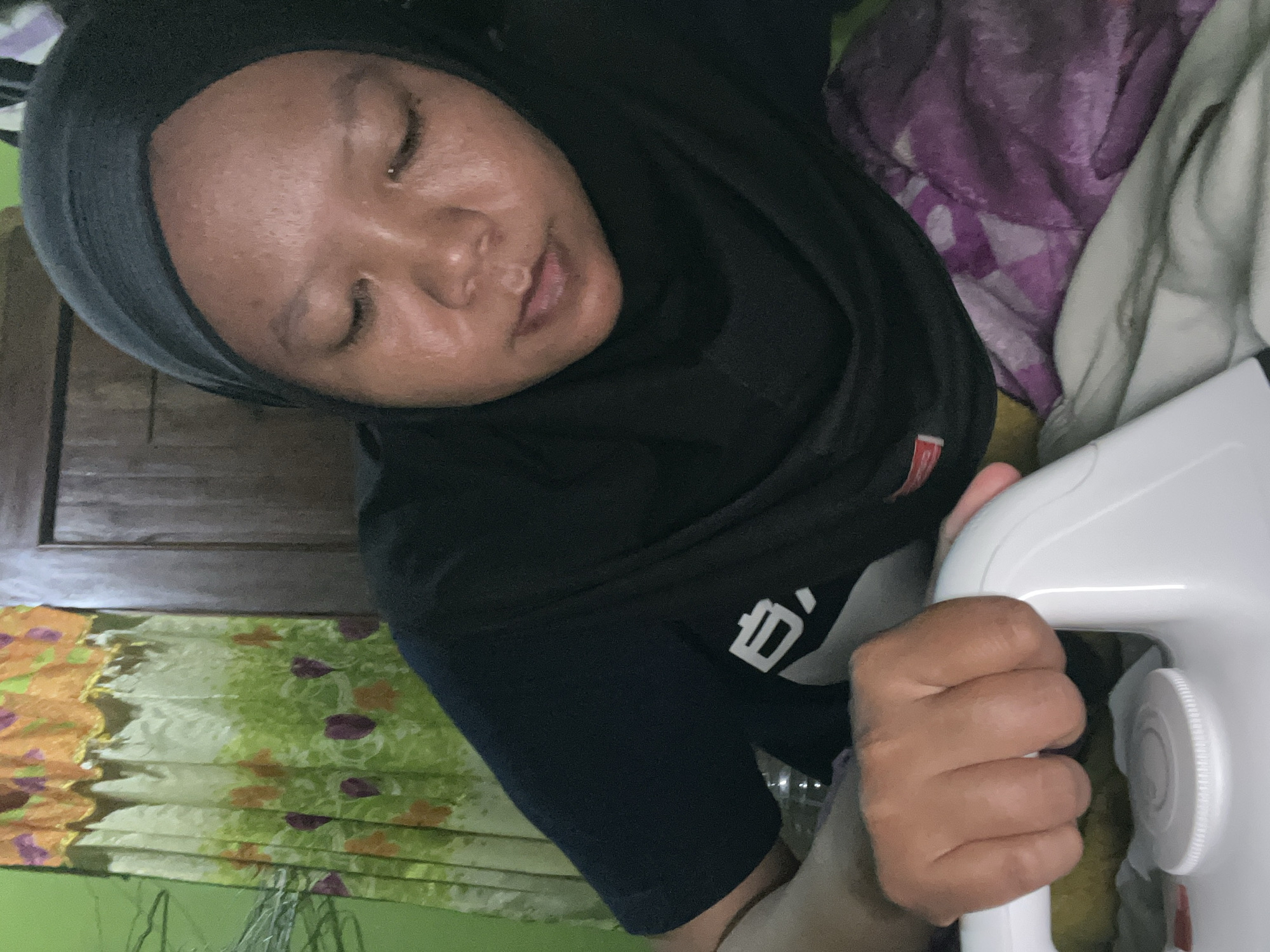 A woman wearing a black hijab and a black t-shirt with a white logo is shown indoors, possibly in the city of Sepat, Indonesia. She is leaning forward and holding a white iron with her right hand. Her eyes are closed, and her facial expression suggests concentration or sleepiness. The background features a curtain with a colorful floral pattern, a wooden closet, and a bed covered with a purple and white patterned blanket.