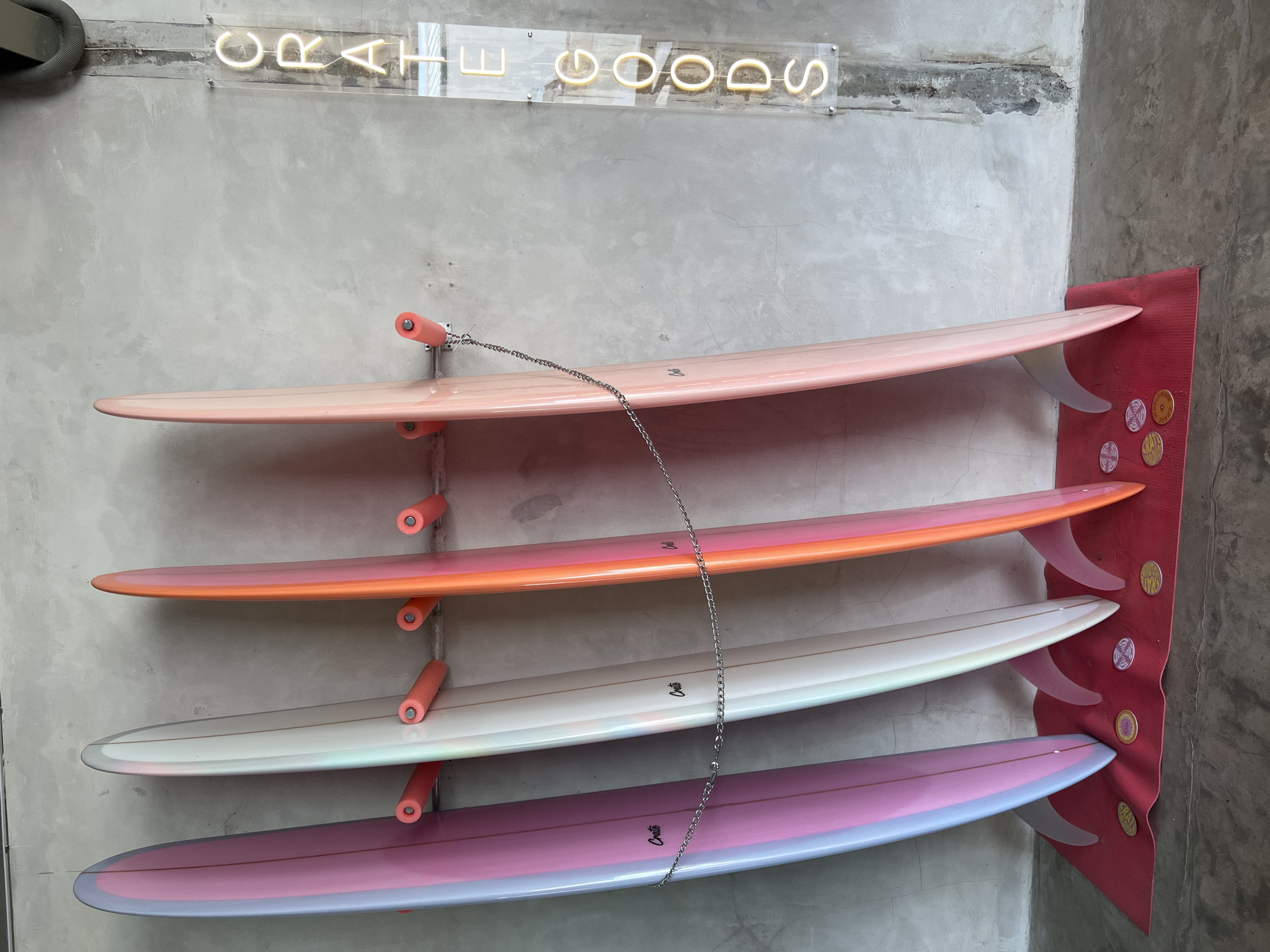 In Mengwi, Indonesia, a row of four surfboards stands upright against a textured concrete wall. The surfboards are arranged from left to right, with the leftmost board displaying a gradient of pink and light blue hues, followed by a surfboard with a white and iridescent sheen, then an orange and pink gradient board, and finally a pale pink board on the far right. Each surfboard has a fin and is supported by orange padded brackets mounted on the wall. A thin metal chain connects the first and last surfboards, with another chain linking the second and third boards. Beneath the surfboards, a red mat is laid out, adorned with several circular, coin-like objects with intricate designs. To the right of the surfboards, a vertical neon sign spells out "CRATE GOODS" in glowing yellow letters against a clear acrylic panel. The floor is a rough concrete surface. There are no people visible in the image.
