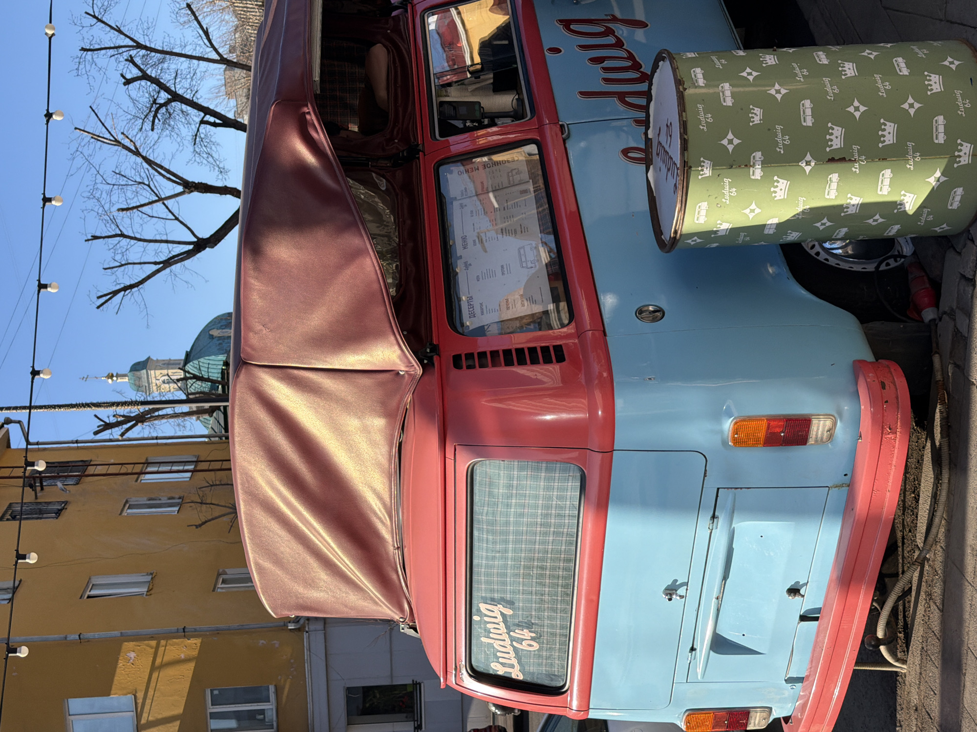 Here is a detailed description of the image:
**Scene**: A vintage, light blue and red Volkswagen van, likely a food truck, parked on a street. The van has "Ludwig 64" written on it, repeated on the canister nearby. A red awning extends from the van's side. A green canister patterned with crowns and small vans, also labeled "Ludwig 64," sits next to the vehicle.
**Location**: The location is Krasnoyarsk, Russia. This is based on the Cyrillic text in the menu displayed in the van's window. In the background, a building with a green dome resembling a church or cathedral is visible. The architecture and style of the surrounding buildings further suggest a European urban environment.
**Environment**: The scene is bathed in sunlight, indicating it's likely daytime. Trees line the street, some with bare branches suggesting either early spring or late autumn. The background includes details of other buildings, windows, and architectural features, giving a context of an urban street scene.