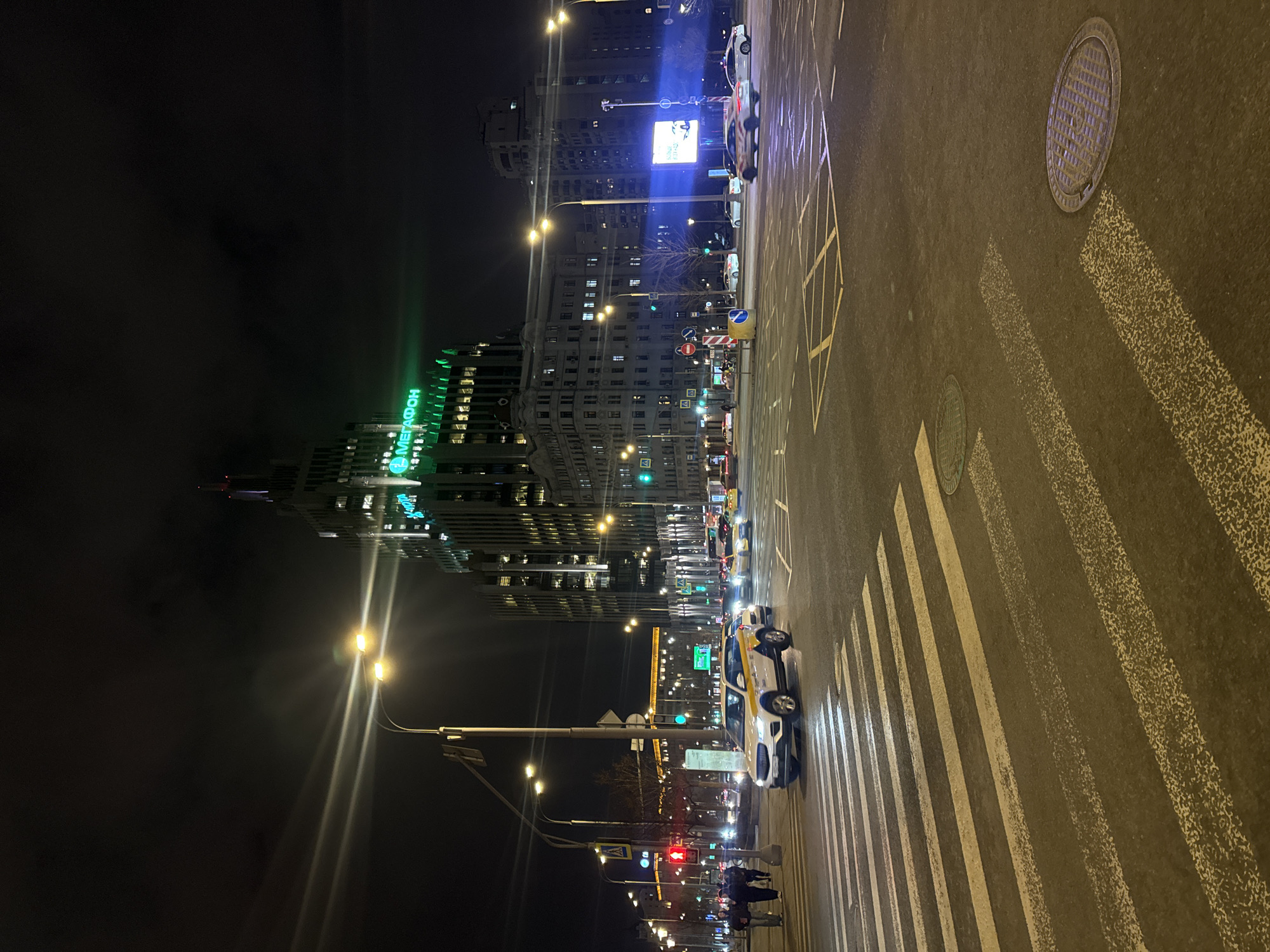 The image depicts a wide urban street intersection at night, illuminated by numerous streetlights and building lights, under a dark, overcast sky. The scene is located at Smolenskaya-Sennaya Square, a prominent intersection on Moscow's Garden Ring, Russia.
Dominating the background is a tall, modern skyscraper, the Smolensky Passage II business center, with a distinctive tiered architectural style, its windows illuminated. A large, bright green neon sign displaying "МЕГАФОН" (MEGAFON) is prominently visible on the upper portion of this building. To the right of this skyscraper, an older, multi-story building with classical architectural elements is visible.
In the foreground, a multi-lane asphalt road stretches across the frame, marked with worn white pedestrian zebra stripes in the immediate foreground and yellow criss-cross "keep clear" markings further back. Two circular metal manhole covers are embedded in the road surface.
Several vehicles are present on the road. A white SUV, possibly a Geely Coolray, with yellow and gray livery (suggesting a taxi or ride-share service) is positioned centrally in the mid-ground, facing right, seemingly stopped or moving slowly across the pedestrian crossing. Multiple other cars, some appearing to be taxis with yellow markings, are visible in the distance across the intersection, some stopped at traffic lights while others show movement with streaking headlights and taillights. A large digital billboard on the right side of the road displays a blue-toned advertisement, likely for a car.
On the left side of the frame, a sidewalk borders the road, where a small group of approximately three to four pedestrians is walking. A pedestrian traffic light displays a red "stop" signal, while vehicle traffic lights further down the road show green. Tall streetlights with multiple fixtures illuminate the scene with bright yellow-white light, creating noticeable lens flares. Various road signs, including regulatory and informational signs, are mounted on poles. Bare trees line the sidewalks, and a blue and white cylindrical barrier is placed on a central road island.