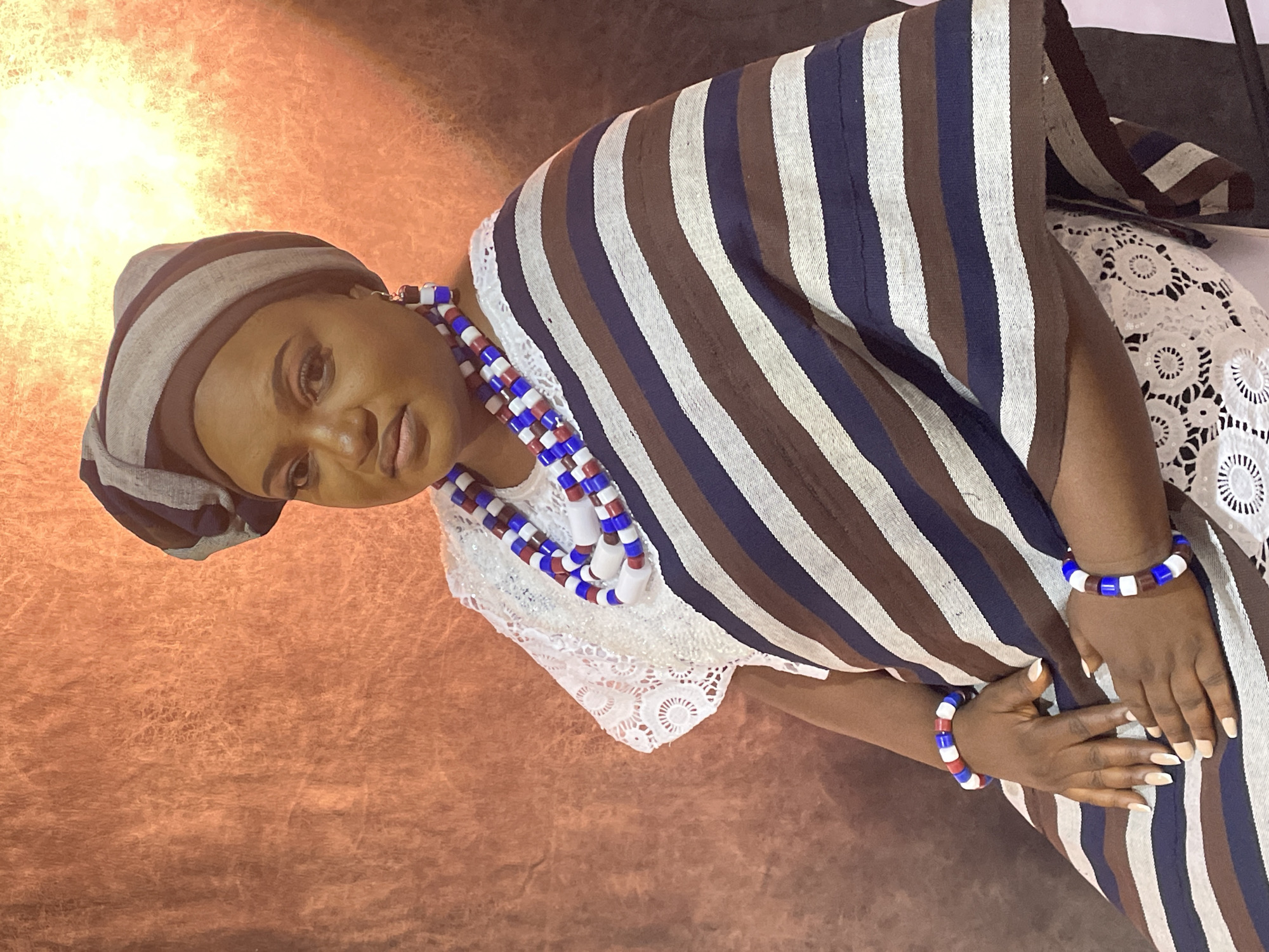 A medium shot depicts a dark-skinned woman with a headwrap, white lace top, and striped shawl in Barakin Akawo, Nigeria. She wears beaded jewelry. The background is a gradient of light to dark brown.