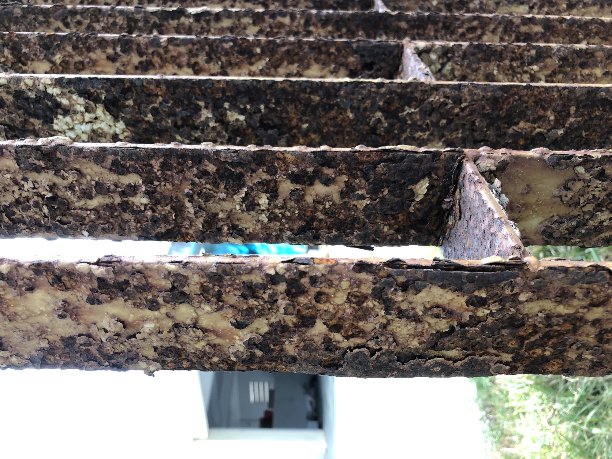 This image depicts a close-up, eye-level view of severely rusted metal bars, likely part of a gate or fence. The bars are vertical and exhibit significant corrosion, with flakes and patches of rust in varying shades of brown, orange, and a lighter, almost cream-colored material. The texture is rough and pitted. In the background, out of focus, is a building with white walls and horizontal window slats, suggesting an outdoor urban environment. There is also some green grass visible in the lower left corner, further indicating an outdoor setting. The overall impression is one of neglect and the effects of weathering on metal. The provided context suggests this scene is located in the city of Đà Nẵng, Vietnam.