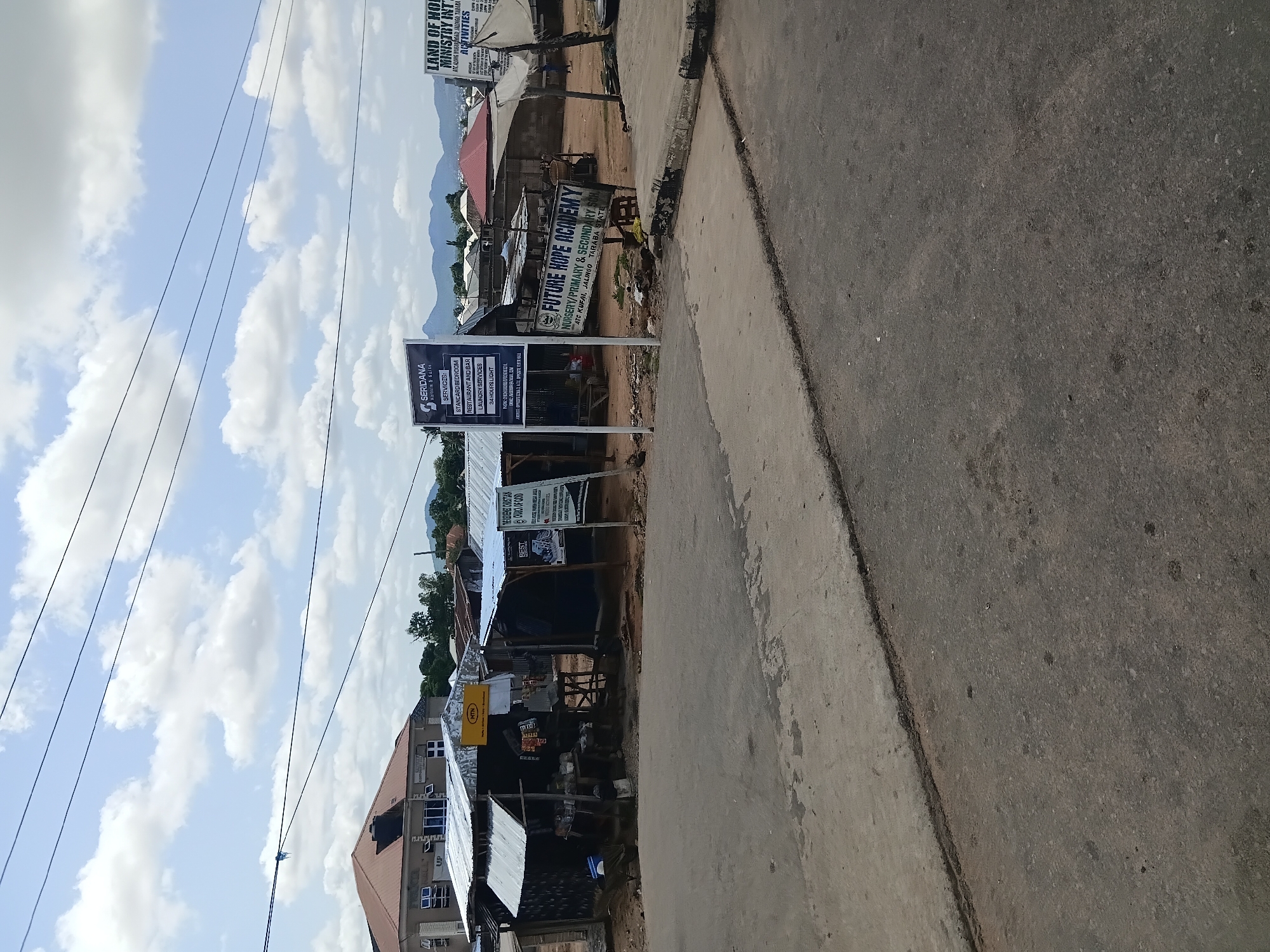 The image captures a street scene in Jalingo, Nigeria, under a partly cloudy sky. In the foreground, a paved road transitions to a dirt or gravel area where several makeshift structures stand. These structures, with corrugated metal roofs and walls, appear to be small businesses or shops. Signs are visible, including one for "SERIDANA Hotels & Suites" listing services like "STANDARD BEDROOM," "RESTAURANT AND BAR," and "LAUNDRY SERVICES." Another sign advertises "FUTURE HOPE ACADEMY NURSERY/PRIMARY & SECONDARY SCH" with the location "ATC KUFAL, JALINGO, TARABA STATE." Further to the right, a sign for "LAND OF HOPE MINISTRY INT'L" is partially obscured. In the background, more buildings and some mountainous terrain can be seen. The overall impression is of a busy, developing urban area.
