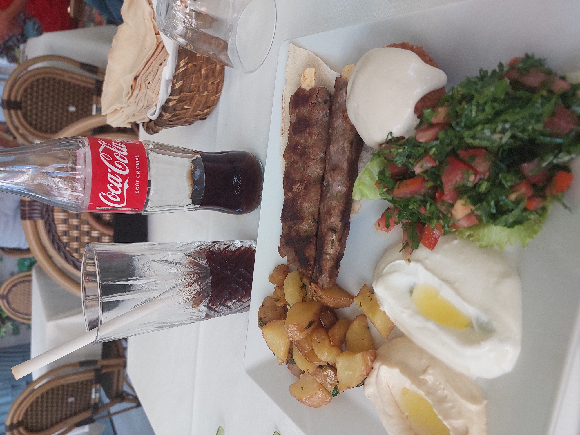 In Antibes, France, at a restaurant, a white tablecloth covers a table. On the table, a white plate holds a meal. The meal consists of two pieces of grilled meat on skewers, served with pita bread. Alongside the meat are roasted potatoes, a serving of hummus, and a creamy white dip, possibly yogurt or a cheese spread. A side of tabbouleh, a salad of finely chopped parsley, tomatoes, mint, onion, and bulgur, is also present, topped with a dollop of the white sauce. A glass with a straw contains a dark beverage, likely cola, next to a glass bottle of Coca-Cola. In the background, wicker chairs and tables suggest an outdoor seating area, with some greenery visible.