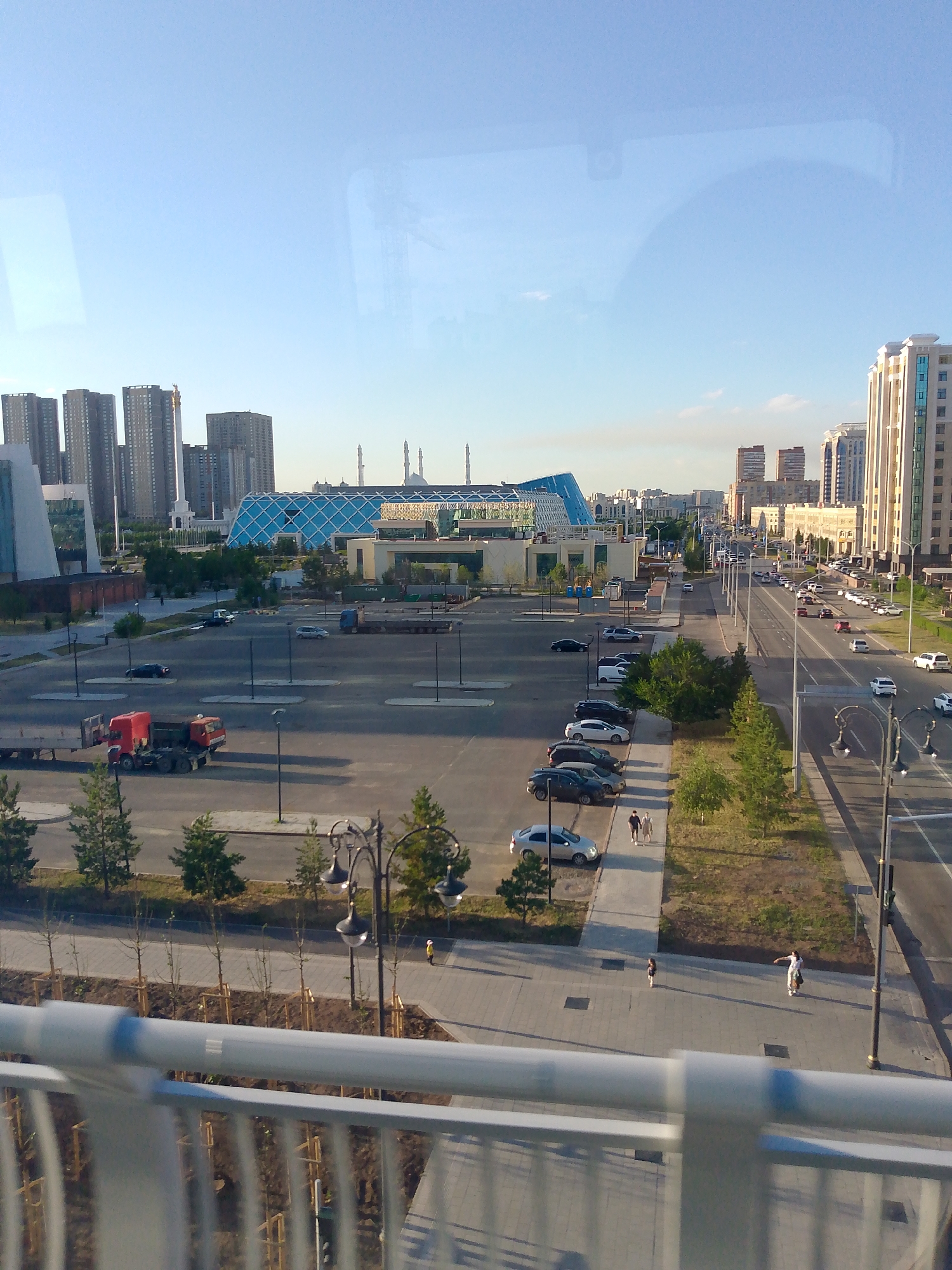 A high-angle, eye-level medium shot captures a panoramic view of a modern city, likely in the Middle East, under a clear, pale blue sky. The foreground features a blurred, light gray railing, suggesting the photo was taken from a bridge or elevated walkway. Beyond the railing, a paved walkway with young trees in neat rows stretches across the mid-ground. A few people are visible on this walkway, including a couple walking together and a child with an adult.
Dominating the mid-ground is a large, empty parking lot with a red truck with a flatbed trailer parked in the lower left corner. Several cars are scattered throughout the lot. Beyond the parking lot, a striking, modern building with a geometric, blue-tiled facade stands out. This building is surrounded by other contemporary structures, including tall residential towers and a white tower topped with a golden spire in the distance.
To the right of the scene, a multi-lane road runs parallel to the parking lot, with numerous cars in motion. Streetlights line both sides of the road. In the background, more buildings and what appear to be minarets can be seen against the horizon. The overall impression is one of urban development and modern architecture bathed in the soft light of what appears to be late afternoon.