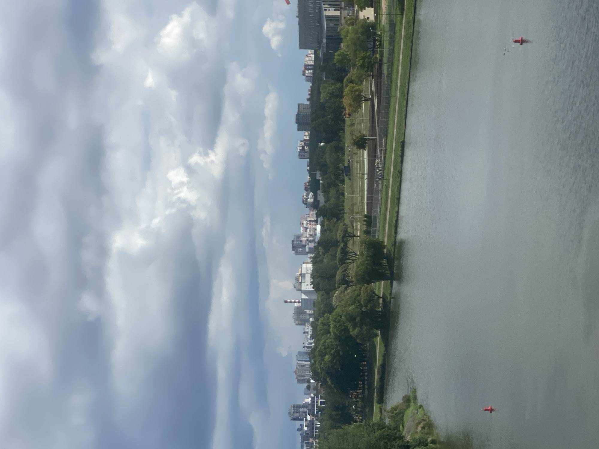 The image captures a wide panoramic view of a cityscape in Minsk, Belarus, on a cloudy day. In the foreground, a tranquil body of water, likely the Svislach River or a related canal, dominates the lower half of the frame. A solitary red buoy marks the water's surface, and a small flock of birds can be seen further in the distance.
The middle ground is characterized by a lush green park area bordering the water. A pathway or road runs through the park, with scattered trees and some distant figures, appearing as small silhouettes, suggesting the presence of people. The park appears well-maintained, with grassy expanses and trees of varying sizes and types.
Beyond the park, the cityscape of Minsk unfolds. A collection of buildings, including residential blocks and industrial structures, are visible on the horizon. Notably, a prominent red and white striped chimney stack stands out among the buildings, indicating an industrial zone. The architecture suggests a mix of modern and older structures. The sky is filled with heavy, dark clouds, casting a muted light over the scene and suggesting overcast or potentially stormy weather. The overall impression is one of a city integrated with green spaces and water bodies, under a dramatic sky.