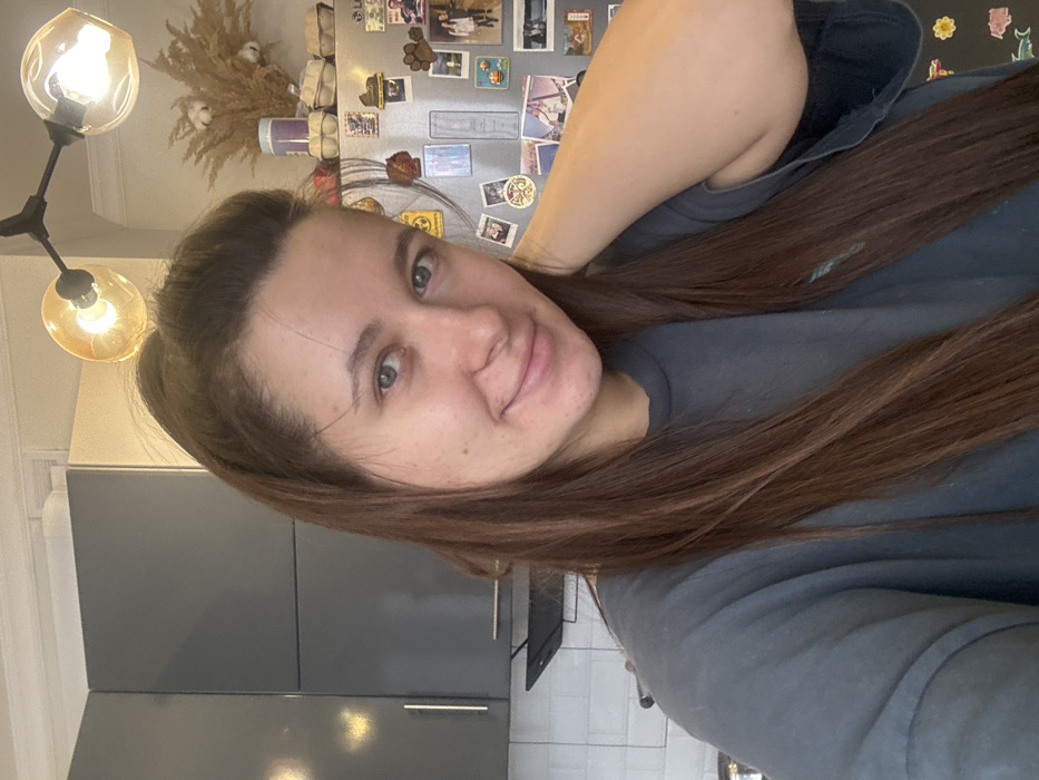 The image displays a female subject in a kitchen located in Odintsovo, Russia. The subject, appearing to be an adult in her 20s or 30s, possesses long, dark brown hair, partially tied back, and has light-colored eyes. She is wearing a dark grey t-shirt and exhibits a slight smile while looking directly at the camera. Her left arm is raised, with the hand positioned behind her head. The background consists of a kitchen interior featuring grey upper cabinets with metallic bar handles on the left. Below these cabinets, a white tiled backsplash is visible, along with a black wall-mounted shelf. To the right, a silver-colored refrigerator is heavily adorned with various magnets, including photographs and symbols. On top of the refrigerator, a decorative arrangement of dried pampas grass and cotton is present, alongside a purple cylindrical container and several light-colored containers resembling egg cartons. Overhead, a black light fixture with two illuminated amber spherical glass shades is suspended from the ceiling.