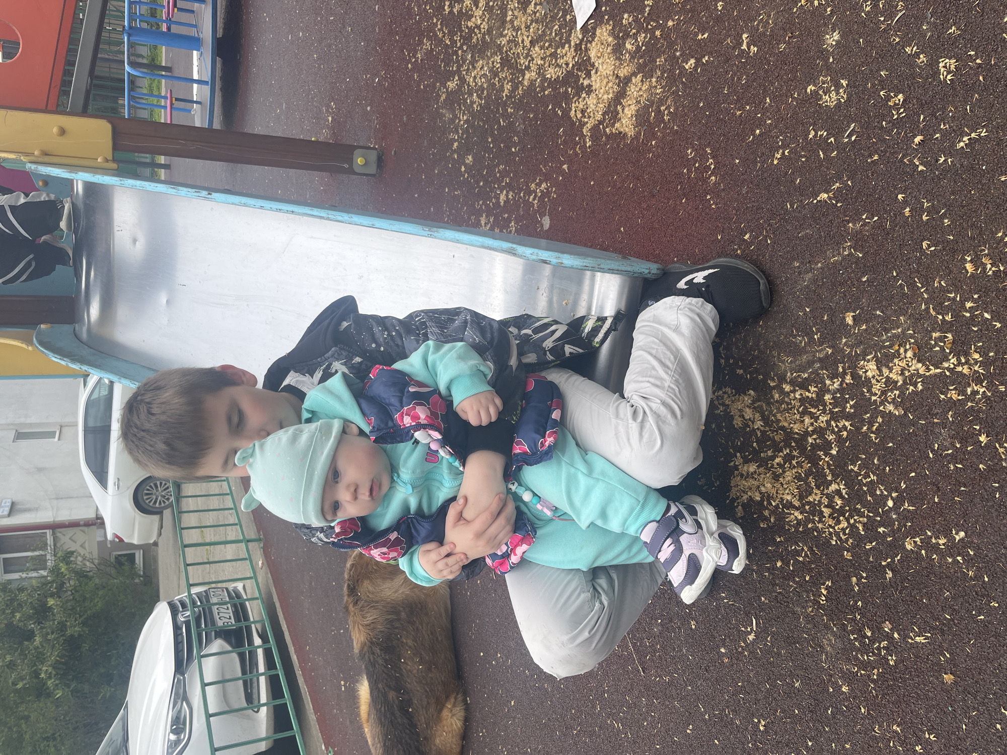 The photo shows a boy, estimated to be around 8-10 years old, holding a baby, estimated to be around 6-12 months old, on a playground. The playground appears to be surfaced with a brown rubberized material and scattered with small pieces of wood debris. A metal slide is visible in the background.
The boy is wearing a dark-colored jacket with a light pattern, white pants, and black shoes with white accents. The baby is wearing a light blue outfit with a patterned jacket and a light blue hat.
A dog is visible in the lower left corner. A white car is partially visible in the background. The license plate on the car reads "B272 HO 01."
Based on the metadata, the location is Vesyoloye, Russia.