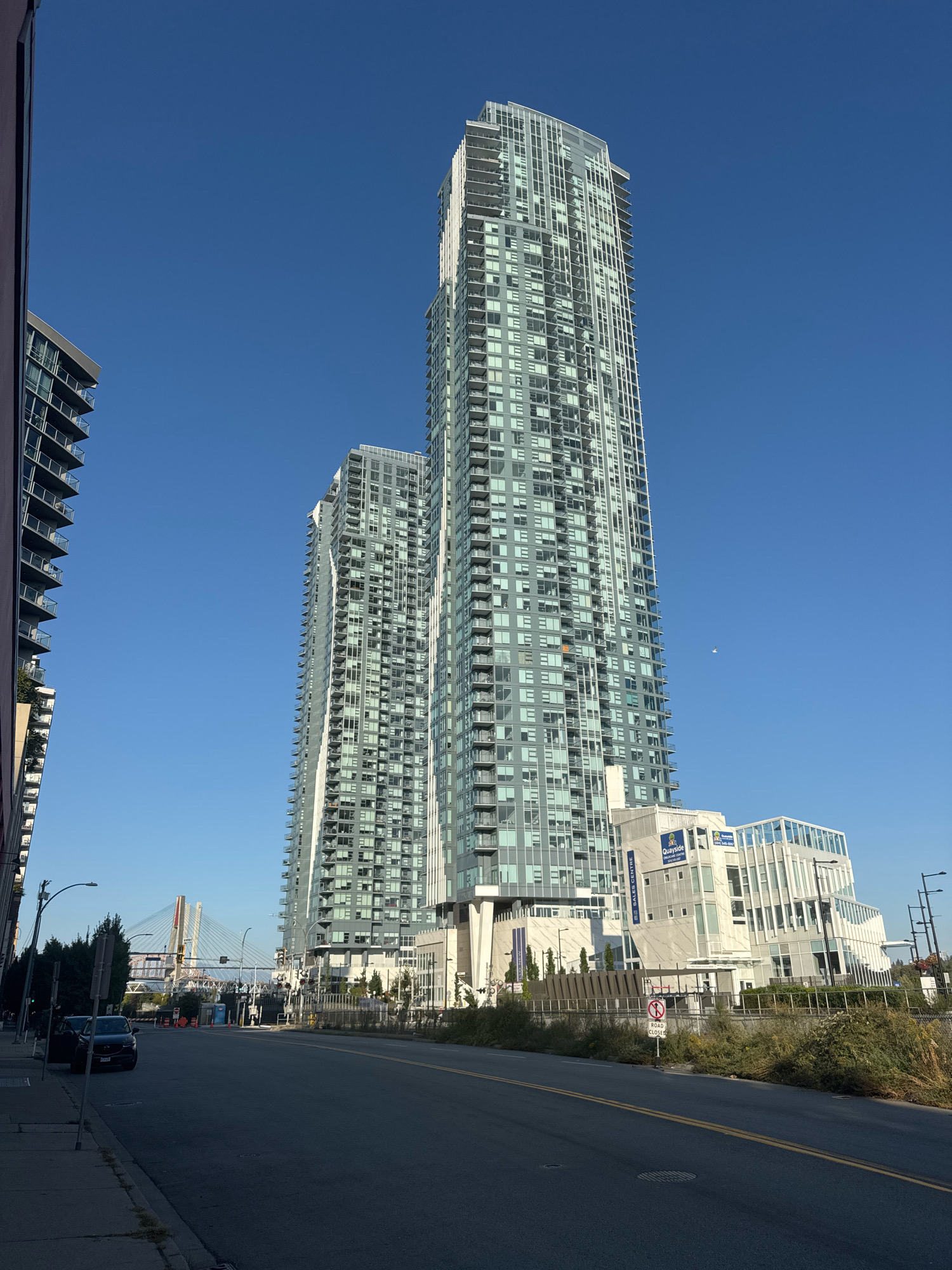 The image shows a street scene in New Westminster, Canada, with modern high-rise buildings dominating the skyline. In the foreground, a dark-colored sedan is parked on the left side of a paved road with yellow lane markings. A sidewalk runs along the left edge of the road, bordered by sparse vegetation. To the right of the road, a chain-link fence encloses an area with overgrown bushes and a "ROAD CLOSED" sign with a "NO U-TURN" symbol.
The focal point of the image is a pair of strikingly contemporary, glass-fronted skyscrapers. The taller building, on the right, features a dynamic, angled design with numerous balconies and a grid of windows. To its left, a slightly shorter, similarly designed skyscraper stands. At the base of the buildings, a modern, multi-story white structure with large glass panels and a distinctive geometric shape is visible, with signage indicating it is the "QUAYSIDE GRAND CENTRE".
In the background, a suspension bridge with white cables is visible spanning a body of water, and a clear blue sky stretches above. The scene appears to be captured during daylight hours, with the sun casting shadows on the road and sidewalk. There are no people clearly visible in the image, and the environment suggests an urban setting undergoing development or with a mix of residential and commercial properties.