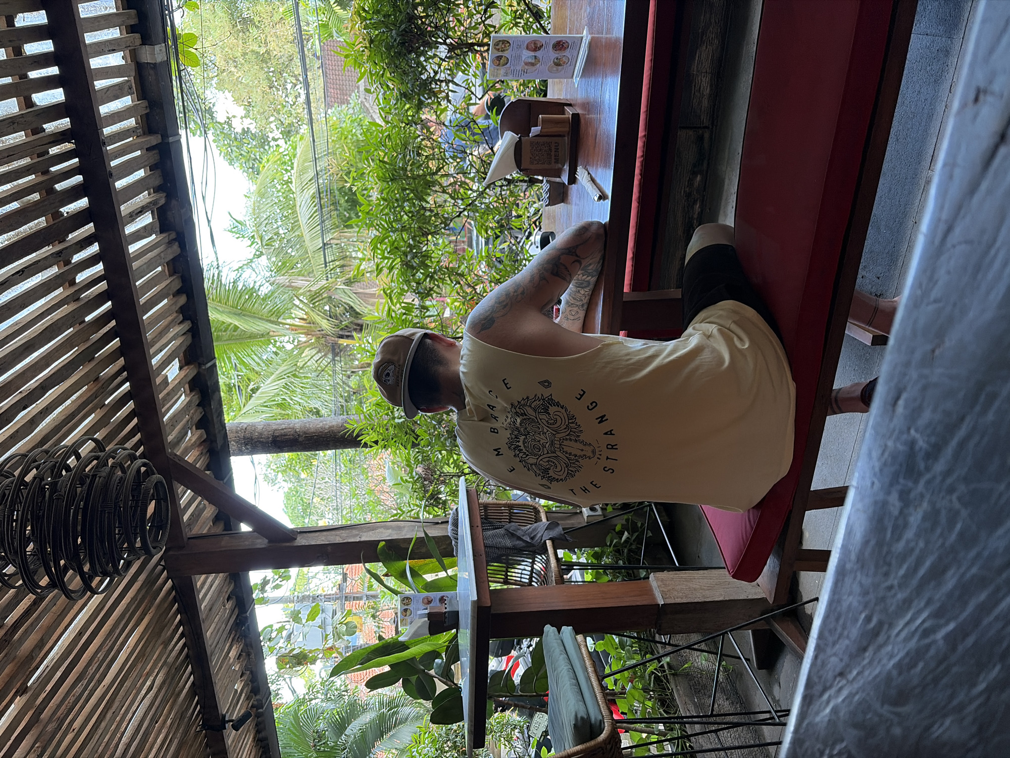 The image shows a person sitting at a wooden table in an outdoor cafe in North Kuta, Indonesia. The person, facing away from the camera, wears a tan baseball cap and a light-colored tank top with a graphic design on the back. They are seated on a red bench. The table is set with a menu, napkins, and cutlery. Lush green foliage and palm trees are visible in the background, suggesting a tropical setting. The cafe appears to have a rustic aesthetic with a wooden roof structure.