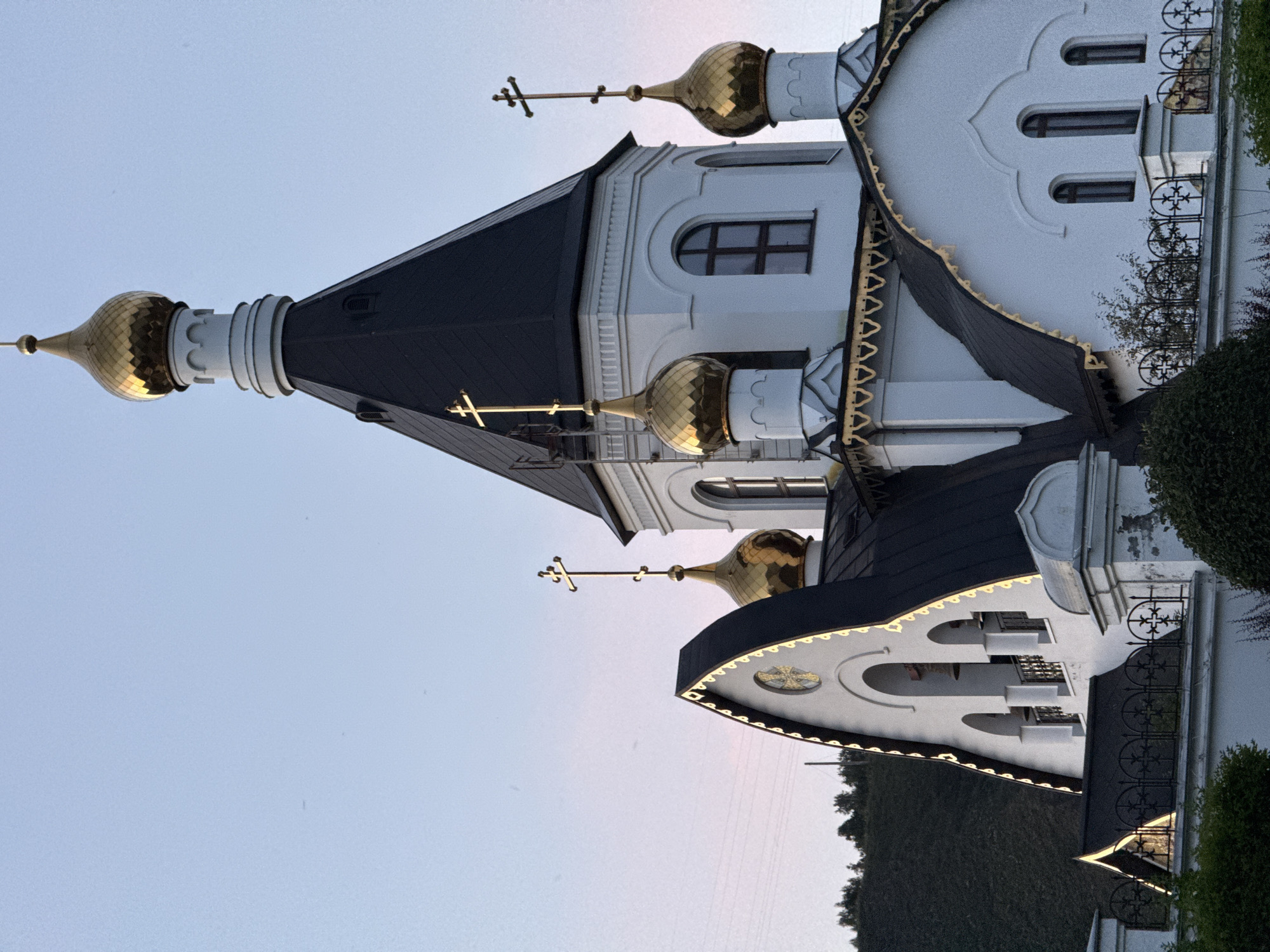 The image shows the exterior of the Transfiguration Cathedral in Krasnoyarsk, Russia. The cathedral features white walls, a dark pitched roof, and golden domes topped with crosses. The sky is a clear, pale blue with a hint of pink near the horizon, suggesting either sunrise or sunset. In the background, a green, tree-covered hill rises behind the cathedral. Power lines are visible in the distance. The overall scene depicts a religious building set against a natural landscape, bathed in soft, natural light.